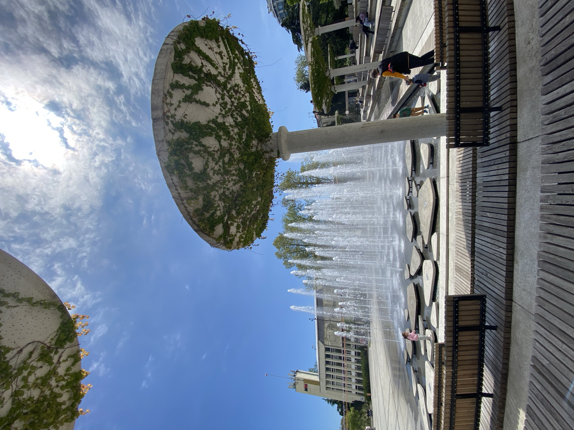 The image depicts a public space in the city of Ялта, Ukraine, featuring a modern fountain. The fountain consists of a large, circular, concrete structure covered in vines, supported by a central pillar. Water jets upward from the fountain's base, creating a visual spectacle. A series of benches line the perimeter of the fountain area. People can be seen walking and sitting near the fountain. The architecture in the background suggests a cityscape with a mix of buildings. The sky is blue with scattered clouds.