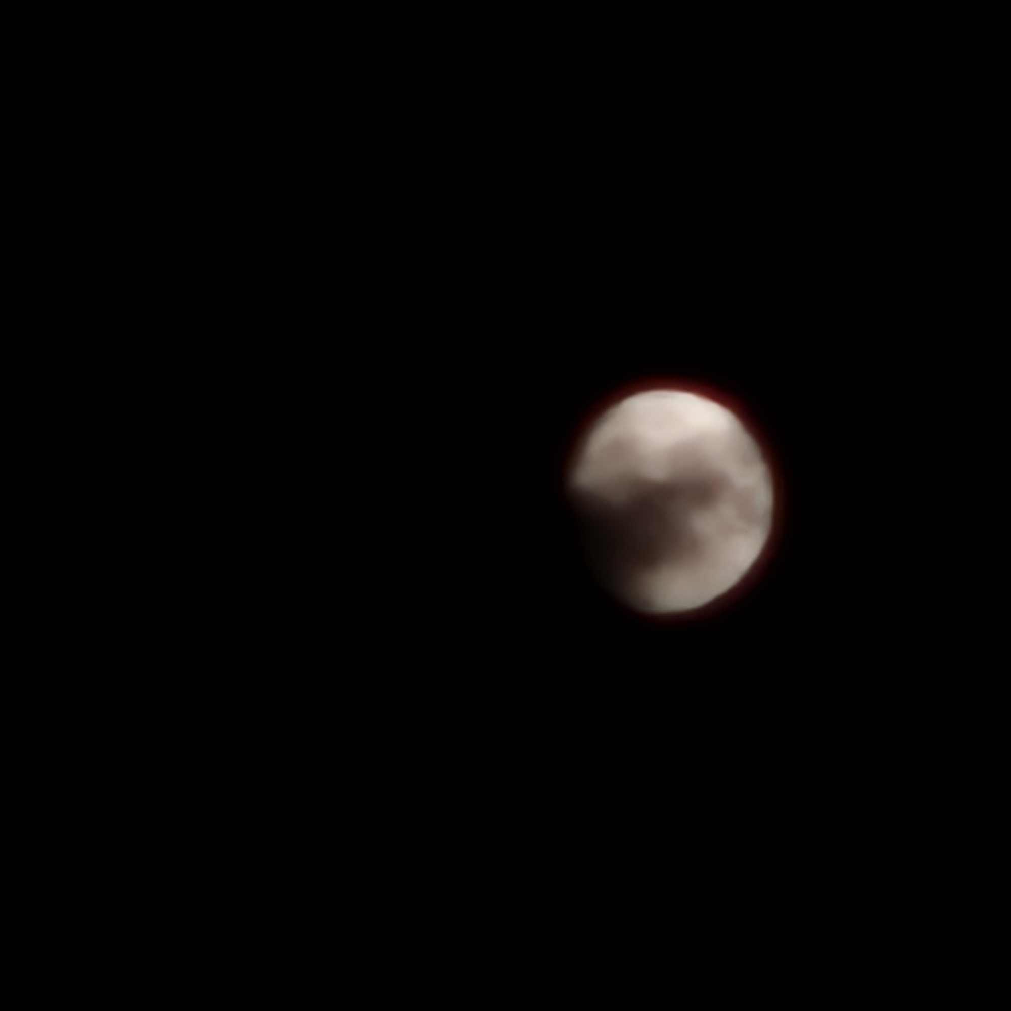 The image is a photograph of a partial lunar eclipse. The moon is visible as a reddish-brown sphere against a black background, with a thin red halo around its edge. Clouds or atmospheric haze are obscuring parts of the moon's surface, giving it a mottled appearance. The image is slightly out of focus, and there is no indication of the city of La Jonquera, Spain.