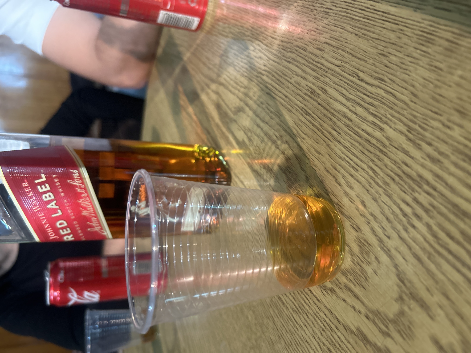 The image shows a wooden table with a plastic cup containing an amber-colored liquid, a bottle of Johnnie Walker Red Label whisky, and a Coca-Cola can. A person is partially visible in the background. The location in Veliko Tarnovo, Bulgaria, cannot be determined from the image.