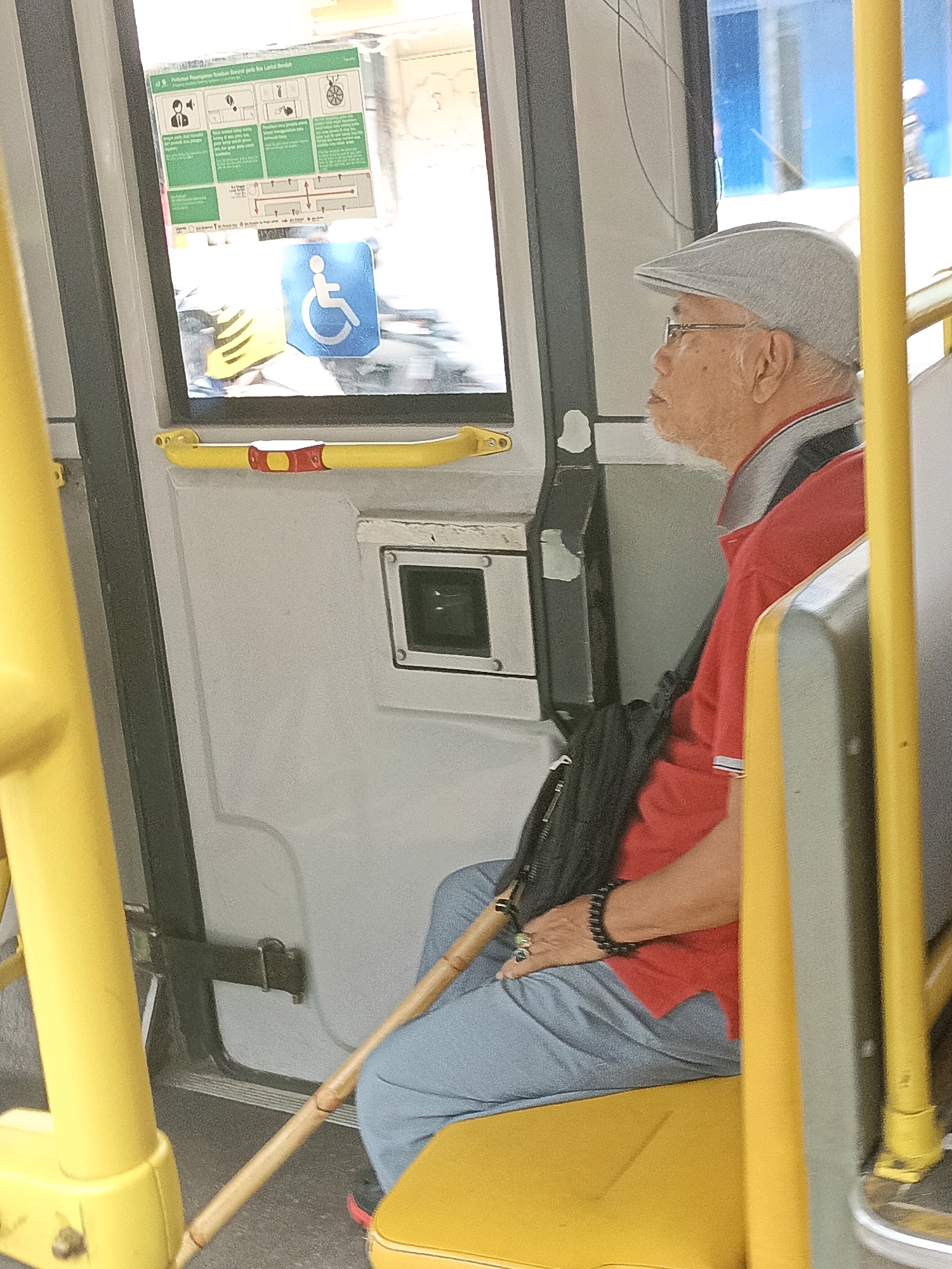 An elderly Asian man wearing a grey flat cap, glasses, and a red polo shirt sits on a yellow bus seat, facing right, with a cane resting against his legs. The man has a white beard and is wearing a black beaded bracelet on his left wrist, with a ring on his index finger. A black bag is slung over his shoulder. The bus interior is visible, including yellow safety poles and a window with safety instructions. A blue wheelchair-accessible symbol is affixed to the window. The scene appears to be inside a public bus, likely in South Jakarta, Indonesia, given the context.