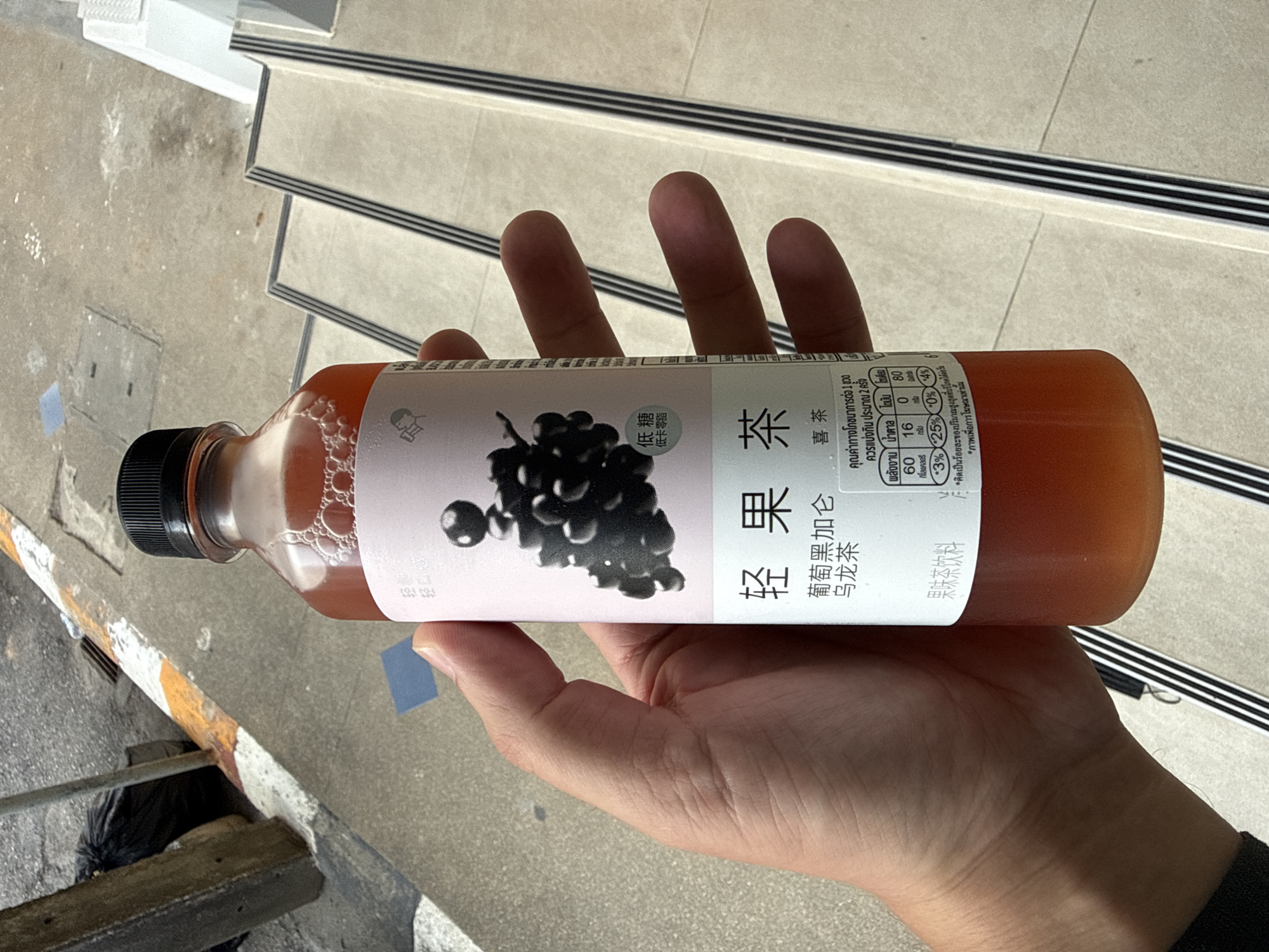 A person's hand holds a clear plastic bottle filled with a reddish-brown beverage, likely tea, with small bubbles visible near the top. The bottle has a black plastic cap and a white label featuring a graphic of dark purple grapes and text in Chinese and Thai characters. The label indicates "low sugar" and mentions "Grape Oolong Tea". A nutritional information chart in Thai is also present on the label. The background shows a concrete surface with a yellow and white striped curb or line, suggesting an outdoor urban or public area. Further back, a set of outdoor stairs with dark treads is visible. The lighting indicates it is daytime, with shadows cast on the ground. The presence of a trash bag and a concrete post in the background hints at a functional, possibly commercial or residential, outdoor space within Phuket City Municipality, Thailand.
