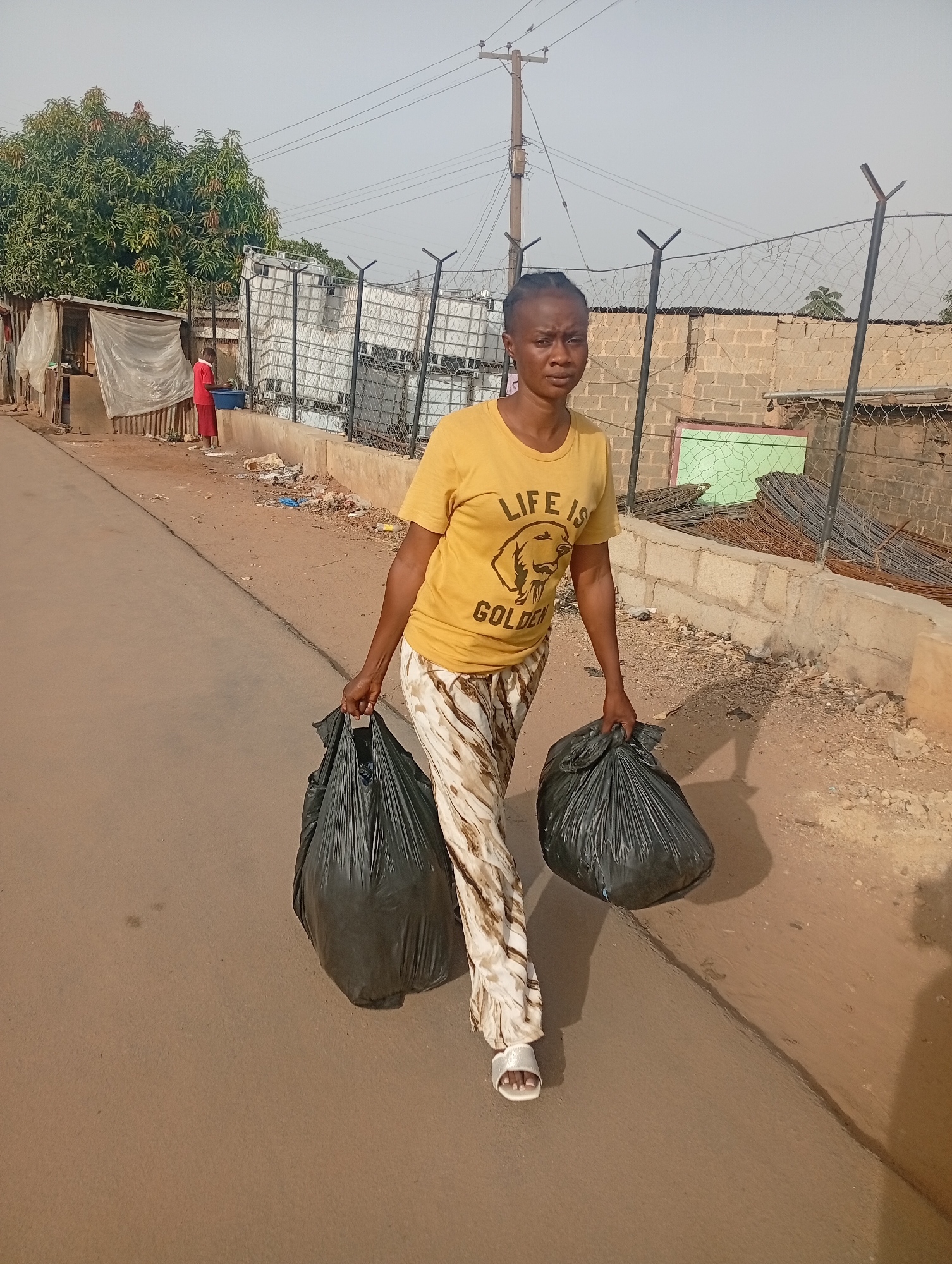 In Jos, Nigeria, a woman walks down a street carrying two black garbage bags, one in each hand. She is wearing a yellow t-shirt with a dog graphic and the words "Life is Golden," as well as patterned pants and white sandals. The street is paved but has dirt and debris along the sides. Behind her, there's a building or structure covered with a tarp, a stack of white containers behind a chain-link fence, and a cinderblock wall topped with fencing and metal bars. Power lines are visible overhead. A person in a red outfit is standing near the tarp-covered structure.