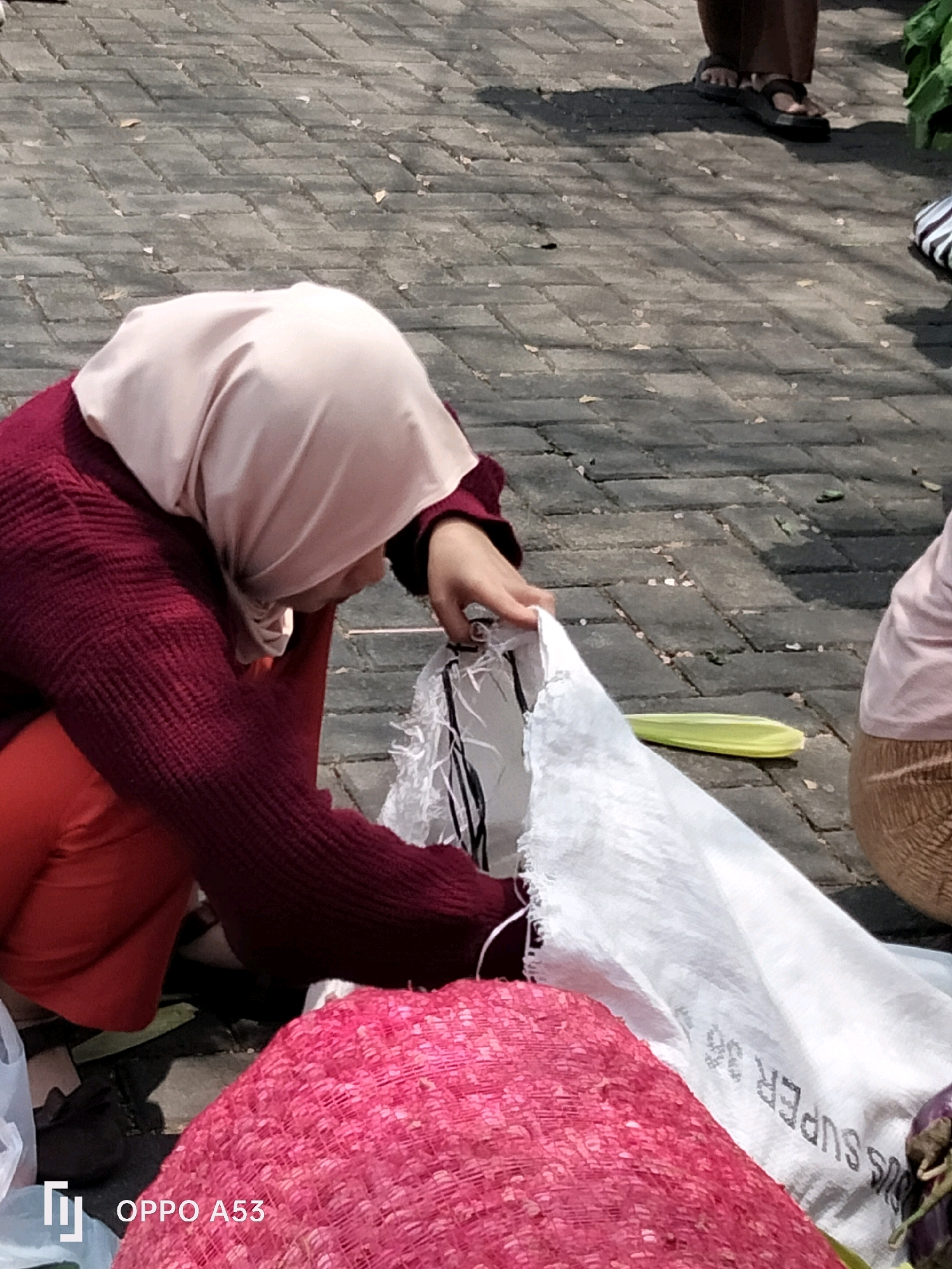 In Kelutan, Indonesia, two individuals are seen in an outdoor setting, likely a market, as indicated by the paved ground and the presence of produce. The foreground features a woman wearing a maroon sweater and red pants, with a light pink hijab. She is crouching and appears to be tending to a large white woven bag. Her right hand is extended towards the opening of the bag. Next to her, a large, bright pink woven object, possibly a basket or a large bundle of items, is prominently displayed.
To the right of the woman, another person, partially visible, is wearing a light pink shirt and a tan skirt. Their lower legs are not visible. Further into the background, another person stands wearing dark brown pants and dark sandals. They are casting a shadow on the paved ground, suggesting a sunny day. The ground is made of interlocking gray pavers, with scattered leaves. A single yellow-green stalk, possibly corn husk, lies on the ground between the woman and the person in the light pink shirt. A portion of a dark-colored vegetable, possibly an eggplant, is visible to the far right, near the person in the tan skirt. The OPPO A53 watermark is present in the lower left corner of the image. The overall impression is of a transactional or agricultural environment.