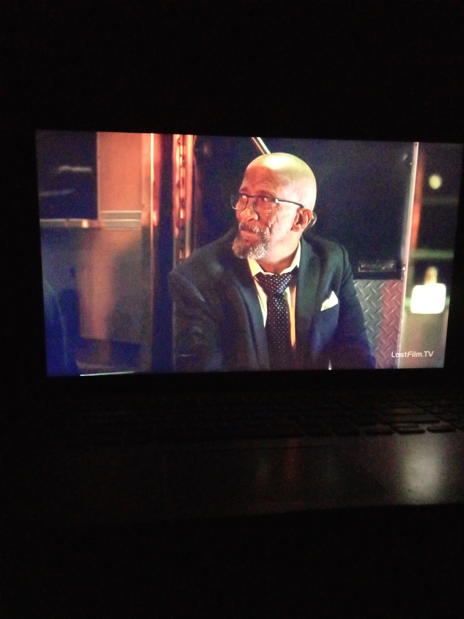 A bald, middle-aged man with glasses and a goatee is seen from the chest up, looking to the left. He is wearing a dark suit jacket, a white shirt, and a black tie with white polka dots. A white pocket square is visible in his jacket pocket. The man appears to be indoors, possibly in a studio or on a set, with a metallic, textured wall behind him. To the right of the man, a white light fixture is visible, and the text "LostFilm.TV" is present in the bottom right corner of the image. The overall lighting is dim, with warm tones suggesting artificial lighting.