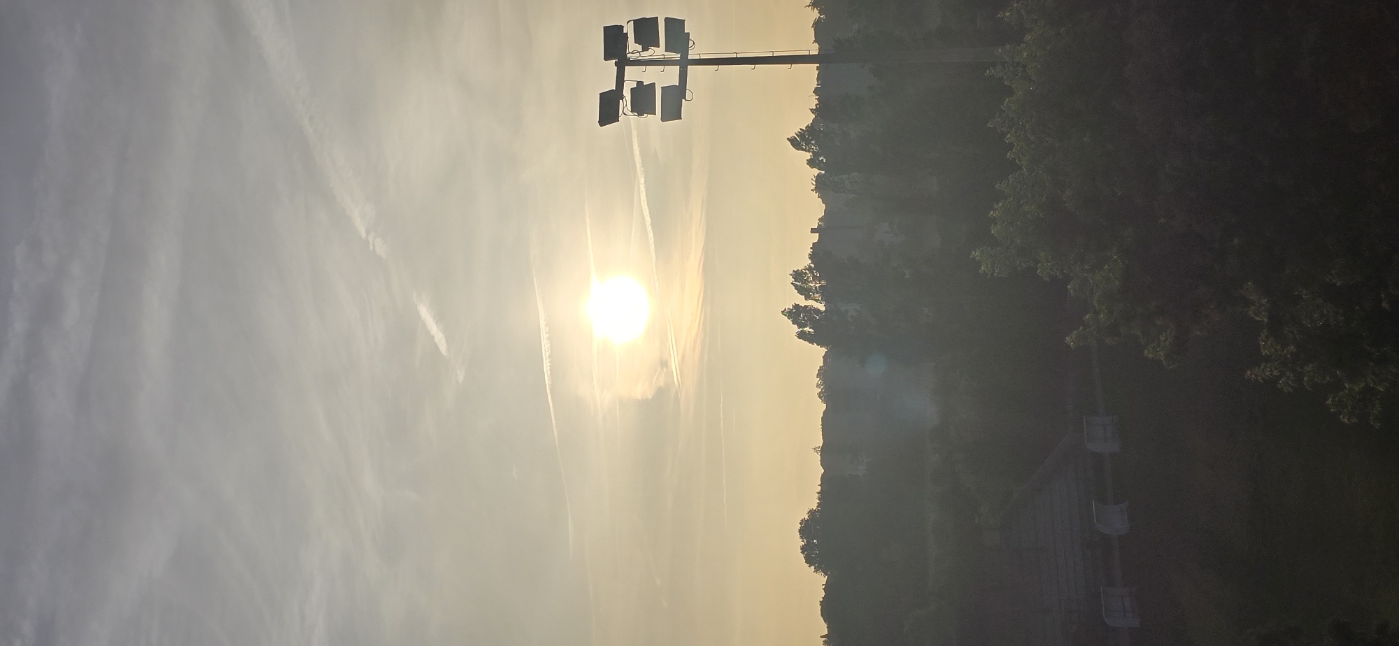 The image depicts a hazy sky at sunrise or sunset, with the sun appearing as a bright, diffused orb. Wispy clouds and contrails are visible in the sky. In the middle ground, a silhouette of trees and houses creates a soft, indistinct landscape. To the right, a tall pole with multiple floodlights is prominently featured, suggesting an outdoor sports facility or stadium. In the foreground and left side of the image, hints of stadium seating and fencing are visible through the haze, further supporting the idea of a sports venue. The overall atmosphere is muted and slightly obscured by atmospheric conditions.
