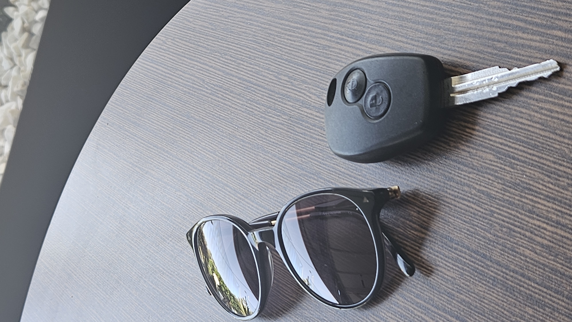 A pair of black sunglasses and a car key are placed on a wooden table. The sunglasses are black with reflective lenses. The car key is black with two buttons and a silver metal blade. The table has a wood grain pattern. The background is blurred and appears to be outdoors, possibly in a cafe or patio setting.