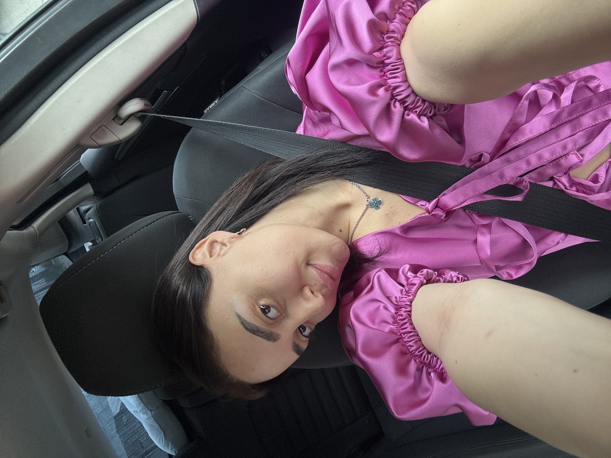 A woman is seated in a car. She is wearing a pink satin top and has a seatbelt fastened across her chest. The car's interior is black, and the woman's head rests against the headrest of the car seat. The exact location in Tbilisi, Georgia, cannot be determined from the image.