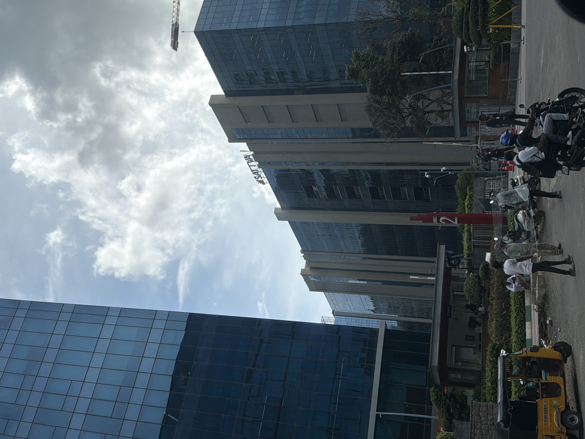 The image depicts the exterior of the Sattva Knowledge City in Hyderabad, India, under a partly cloudy sky. Several modern, multi-story buildings with glass facades dominate the scene. One building on the left is a tall, dark blue glass skyscraper. To its right, a series of connected buildings feature a mix of glass and concrete exteriors, with the "SATTVA" logo visible on the top of one section. A construction crane is visible in the distance above the buildings. In the foreground, there is a street with an auto-rickshaw, several motorcycles, and pedestrians. Two men in white shirts and dark pants are standing near the auto-rickshaw. Other individuals, including security personnel in uniforms, are visible near building entrances and a gate labeled "GATE 2". Landscaping with trees and bushes surrounds the buildings. The overall impression is of a bustling corporate campus in an urban setting.