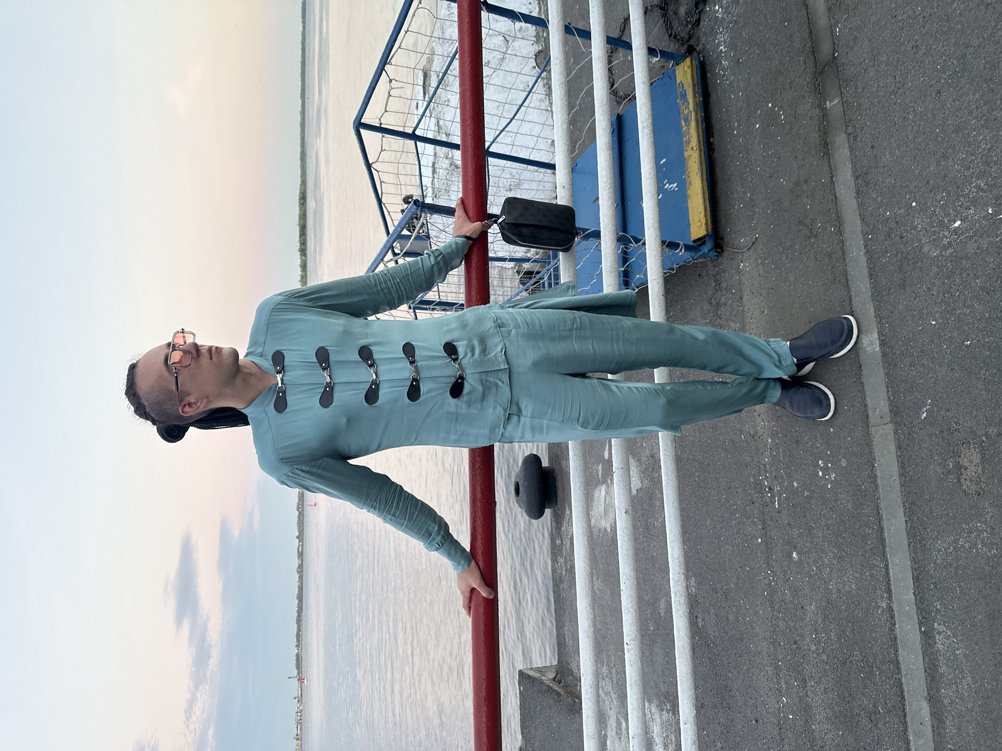 A full-body, eye-level outdoor shot shows a person with dark hair styled in braids, wearing a teal-colored outfit with a unique button closure and square-shaped, rose-tinted sunglasses. They stand on a paved area near a body of water, with their left hand resting on a red railing. A black pouch hangs from the railing near their hand. The background features a pale sky with wispy clouds, a calm body of water, and a distant shoreline with buildings. The overall impression is of a leisurely moment captured against a serene natural backdrop. This scene is likely on the embankment of the Volga River in Saratov, Russia, possibly near the Saratov Bridge or the city's waterfront, given the presence of a body of water and the characteristic flat landscape visible in the distance.