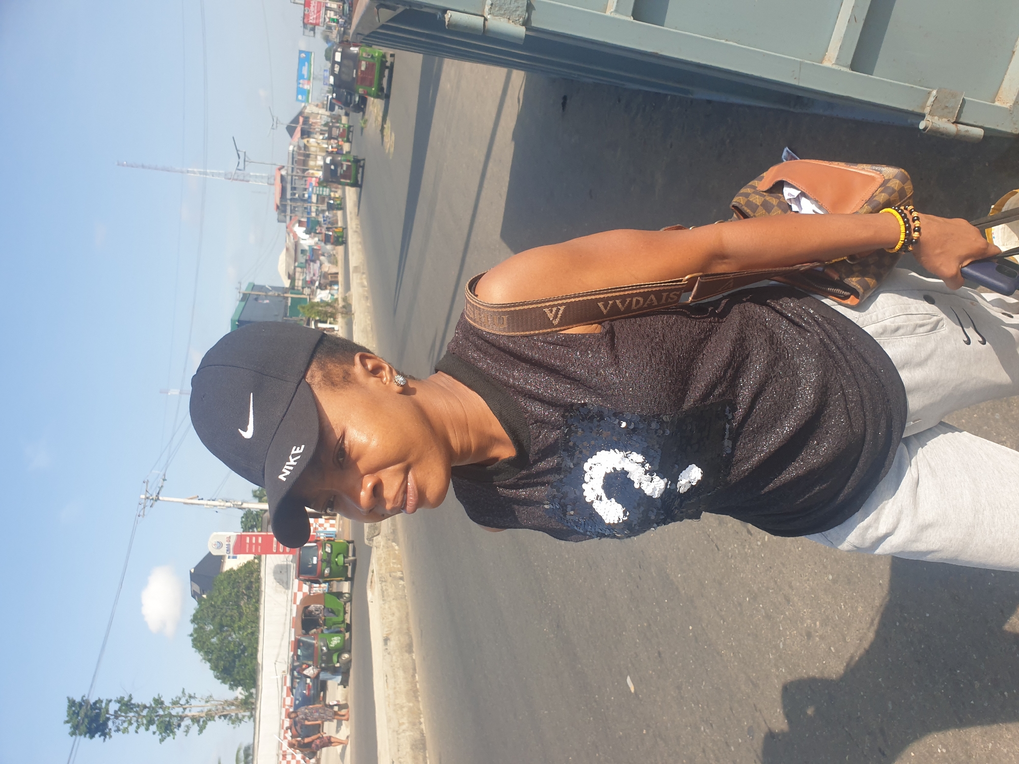 A woman wearing a black Nike baseball cap, a black tank top with white sequin accents, and light gray sweatpants stands on the side of a road in Aba, Abia, Nigeria. She has a brown handbag slung over her shoulder and is holding a blue mobile phone. The background includes a paved road, buildings, and several green tricycles ("keke") a common form of transportation in the city. Overhead power lines and a tall antenna tower are visible. The time of day appears to be sunny daylight.