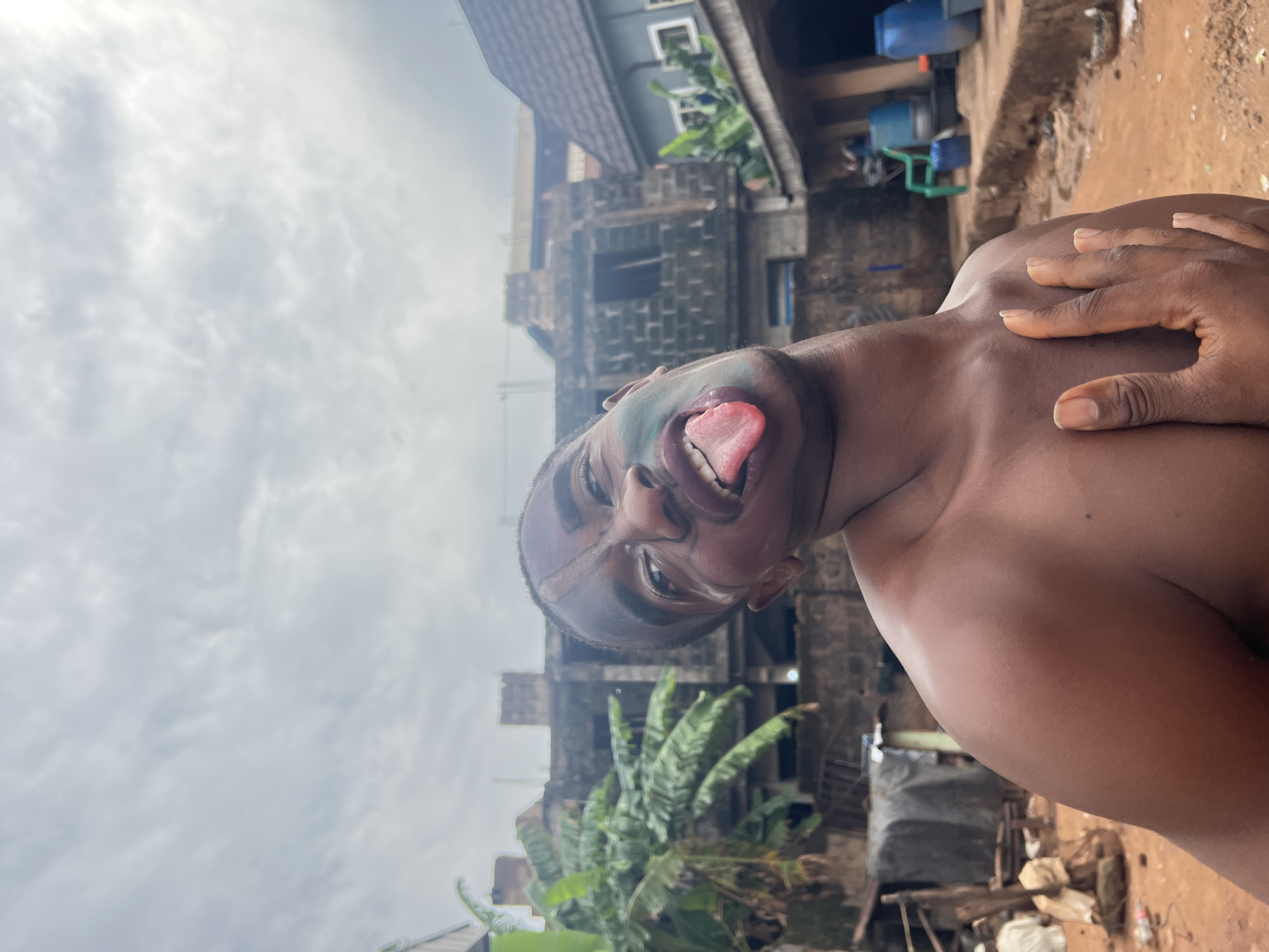A man with face paint, including a line down the center of his forehead and under his eyes, is sticking out his tongue. His chest and shoulder are bare. One hand rests on his shoulder. He is outdoors in what appears to be Onitsha, Nigeria. The background includes houses and banana trees, with a cloudy sky above. There is loose debris on the ground.