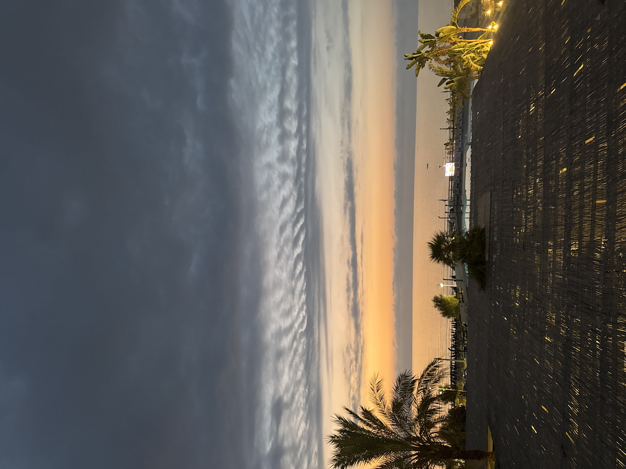 The image captures a twilight scene at a resort or hotel overlooking the sea in Capaci, Italy. In the foreground, a dark, thatched roof structure is visible, with sunlight reflecting off it in long, thin streaks, suggesting it's a covered patio or a rooftop area. Beyond the roof, a swimming pool area is illuminated, with lounge chairs lined up and palm trees surrounding it. The sea stretches out to the horizon, bathed in the warm glow of the setting sun, which is peeking through the clouds. The sky is a dramatic mix of dark grey clouds in the upper portion and lighter, colorful hues of orange and pink near the horizon. Several small boats are visible on the water, and distant lights suggest buildings or other structures along the coastline. The overall atmosphere is serene and peaceful, indicative of the end of a day in a coastal resort town.