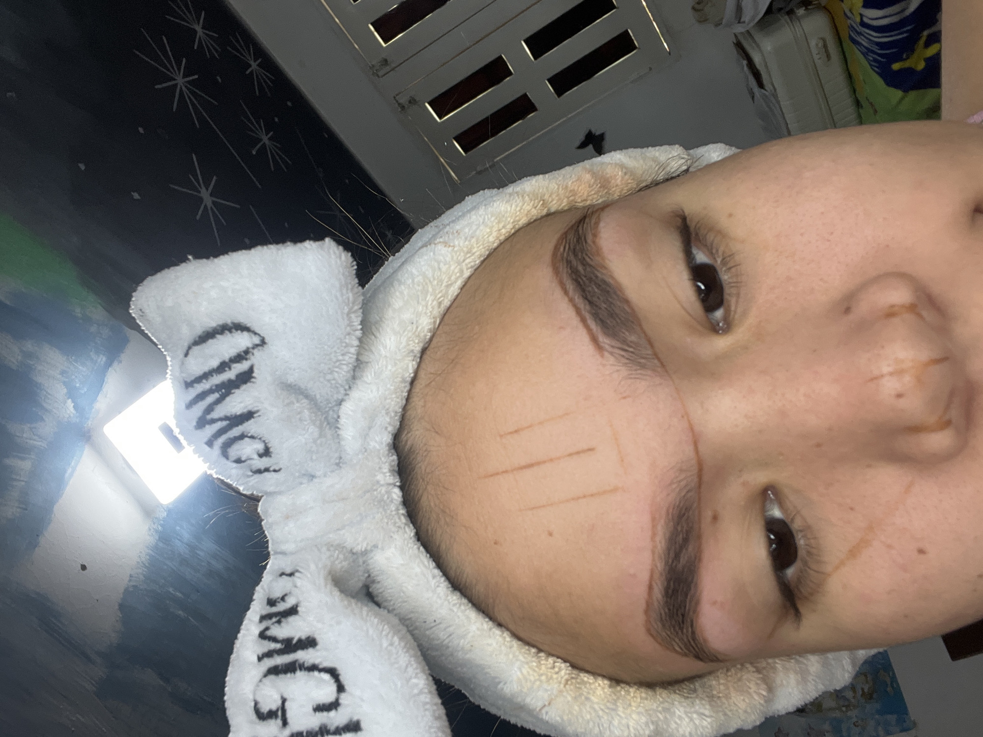 A close-up, head-on shot captures a person's face and forehead. The individual is wearing a white fuzzy headband with black lettering that appears to read "OMG." The headband is adorned with two large, fluffy white "bunny ears" on top. The person's eyebrows have been outlined with a light brown pencil, with additional vertical and horizontal lines drawn on the forehead, suggesting a markup for a procedure. There are also light brown markings on the cheeks and nose. The background is dimly lit, with a bright light source visible in the upper center of the frame. To the right, a ventilation grille and stacked luggage can be seen. The image appears to have been taken indoors, likely in a residential setting in Juan Griego, Venezuela.
