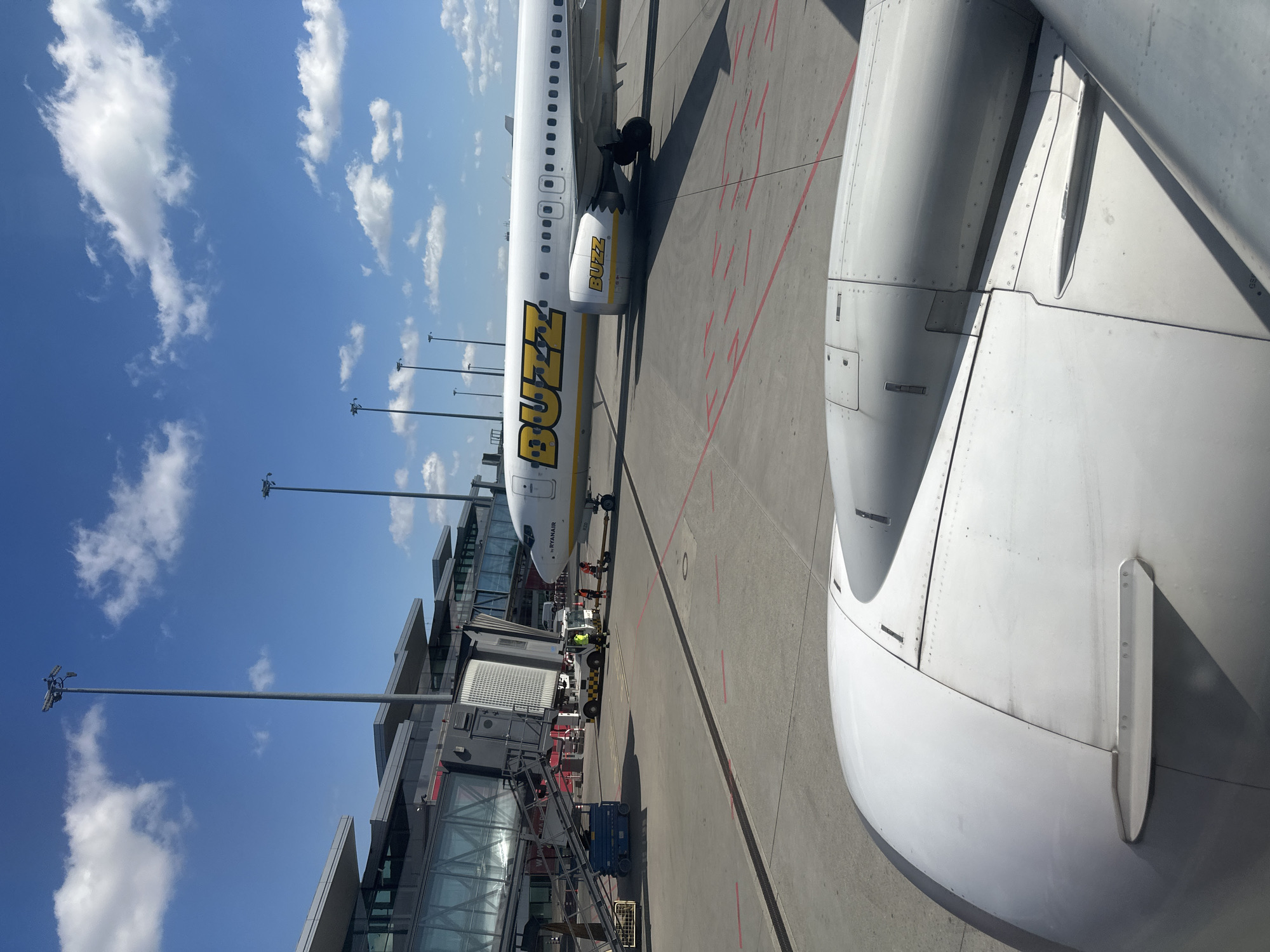 The image, taken from inside an aircraft, shows a Buzz airline plane parked at a gate, likely at Wrocław Airport in Poland given Buzz's presence there. The exterior of the plane, clearly marked with "BUZZ" in large yellow letters, is visible alongside ground crew activity. An airport terminal with glass walls and connecting walkways stands adjacent to the plane. The sky is blue with scattered clouds. In the foreground is a portion of the wing of the aircraft the photo was taken from.