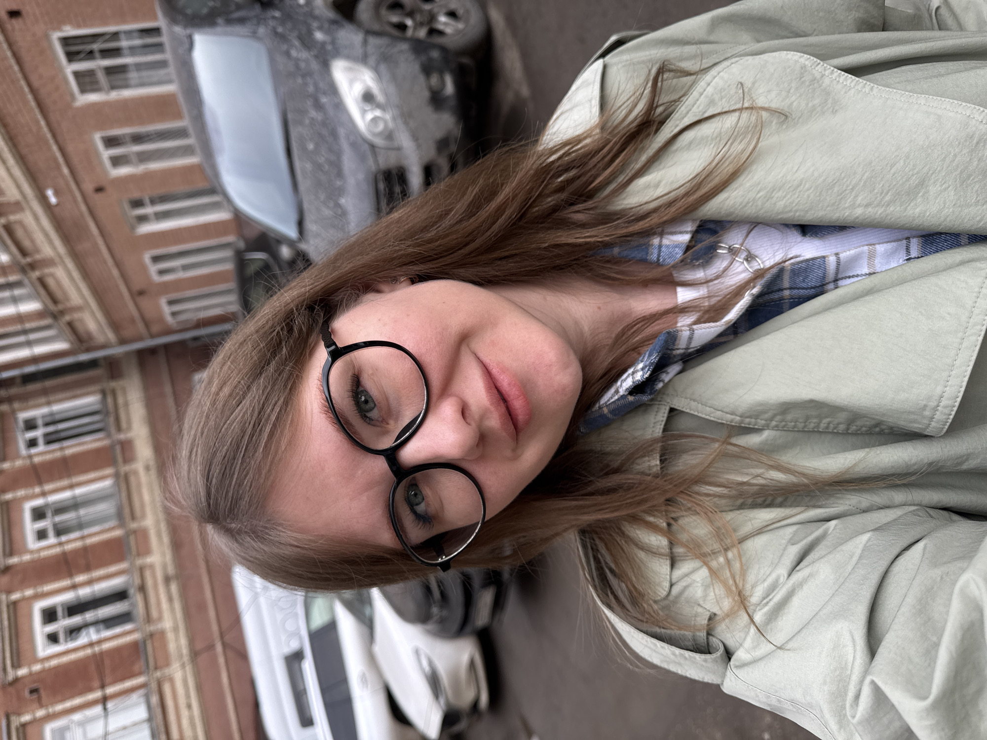 A female individual, approximately 25-35 years old, is depicted in a close-up, chest-up shot in an urban outdoor setting in Perm, Russia. She has shoulder-length light brown hair and wears black-rimmed circular glasses. Her attire consists of a light sage-green trench coat worn over a blue and white plaid shirt and a white undershirt, with a silver-colored chain necklace visible. She looks directly at the camera with a slight smile. The background reveals parked vehicles, including a white van, two light-colored sedans, and a dark-colored SUV heavily coated in dirt or dust. Behind the vehicles, a multi-story building constructed of reddish-brown brick with multiple white-framed windows is visible. The ground is dark pavement.