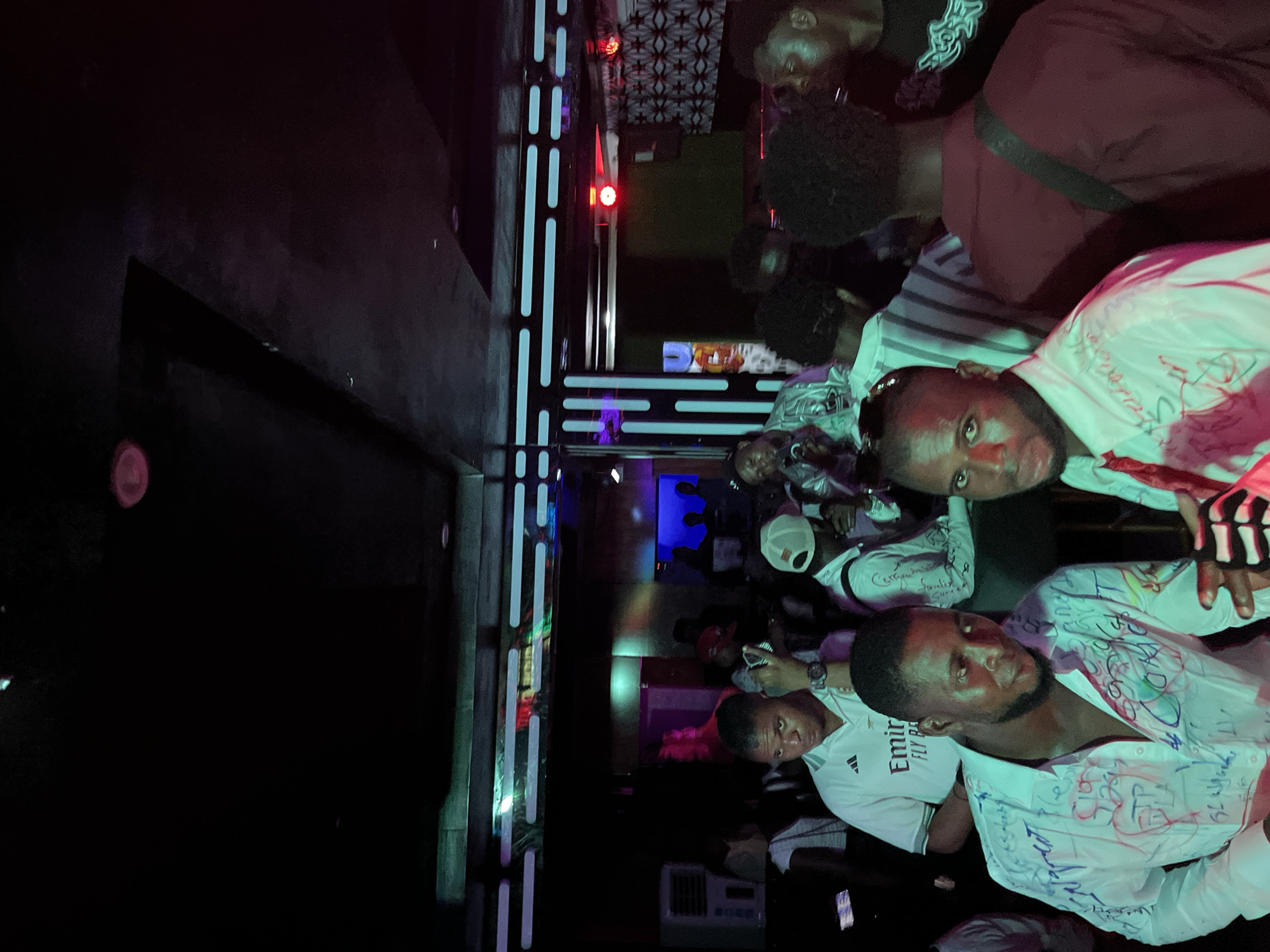 In the dimly lit interior of a nightclub, a group of young men are gathered, some engaging with each other. The scene is characterized by vibrant neon lights illuminating the space, with a large, white air conditioning unit visible on the left. One man in the foreground wears a white shirt with a distinctive collar and appears to be speaking. Another man to his left sports a white shirt adorned with colorful graffiti. The background reveals a bar area with patrons and a DJ booth, where people are actively interacting and enjoying the atmosphere.