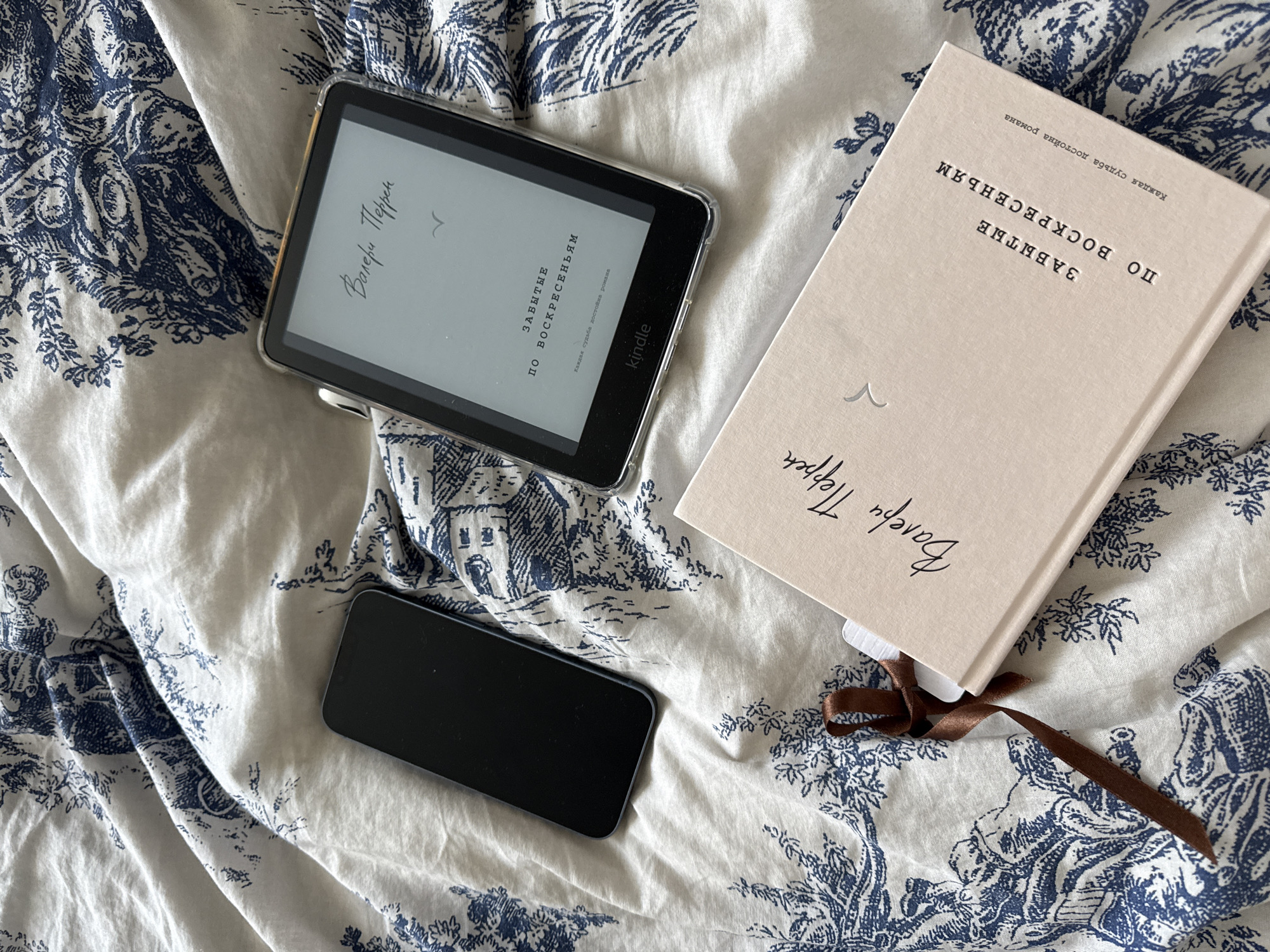 A flat lay image shows a Kindle e-reader and a smartphone on a bed with a blue and white toile-style patterned duvet cover. The Kindle is open and displays the title page of a book in Russian, with the author's name "Валерий Перрен" visible in handwritten script. Below this, the title "ЗАБЫТЫЕ ПО ВОСКРЕСЕНЬЯМ" (Forgotten on Sundays) is printed in text. The bottom of the Kindle screen shows the "Kindle" logo. To the left of the Kindle is a black smartphone, also lying flat. In the lower half of the image, a physical book with a pale pink cover is placed at an angle. The book also displays the author's name "Валерий Перрен" and the title "ЗАБЫТЫЕ ПО ВОСКРЕСЕНЬЯМ" in Russian. A dark brown ribbon bookmark is tied to the book. The background is a rumpled duvet cover with a recurring pattern of blue pastoral scenes and floral motifs. There are no people or animals present in the image. The scene is indoors and appears to be a domestic setting. The image does not provide any specific information about the city of Krasnodar, Russia.