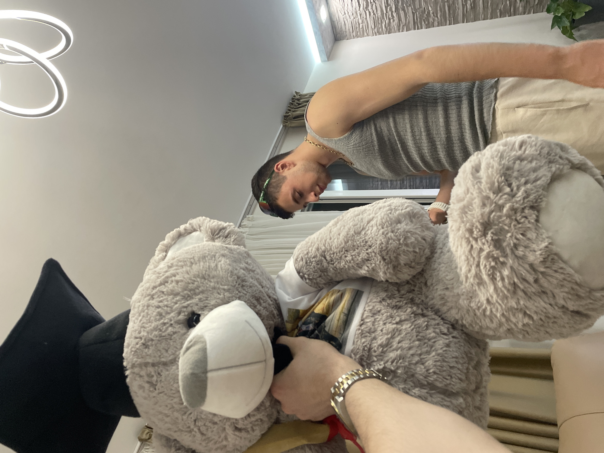 A man wearing a gray tank top, tan shorts, gold chain, and sunglasses sits near a window. He looks downward in the direction of a person's arm and hand that are holding a large, gray, graduation-themed teddy bear. The background includes a white ceiling, modern ring-shaped light fixture, and a wall with stone-like texture. The exact indoor location in Plovdiv, Bulgaria is undeterminable based on the image.