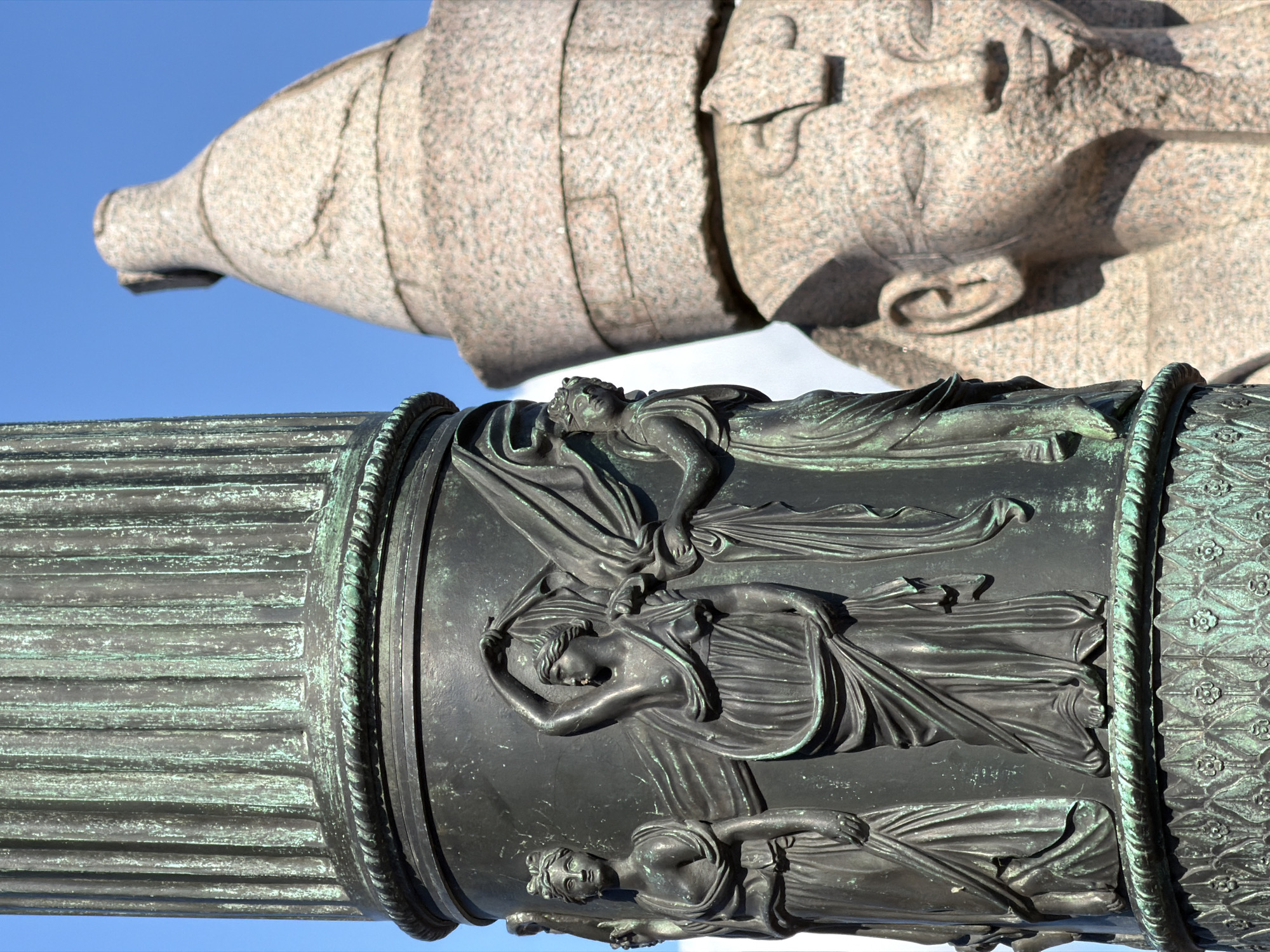 The image presents a close-up view of two distinct monumental sculptural elements set against a clear blue sky, characteristic of a grand public monument in Saint Petersburg, Russia.
In the foreground, a large section of a dark, patinated bronze column dominates the lower half of the frame. This column features intricate bas-relief carvings of several draped female figures, rendered in a classical style. These figures, with their serene expressions and flowing garments, are depicted in various poses, suggesting allegorical representations such as muses or virtues. The column surface shows a rich greenish-black patina, indicative of age and exposure, with fluting on the left and a decorative banded pattern on the right.
Above and behind the bronze column, a large, rough-hewn stone sculpture, likely granite, is partially visible. This element, appearing to be part of a larger figure, shows a stylized human head and upper torso area. Details like an ear, nose, and part of a mouth are discernible, carved from the light, speckled pinkish-beige stone.
The bright blue sky and prominent shadows cast on both sculptures suggest a sunny day, likely midday or early afternoon. No specific activity is taking place as these are static artistic structures. The monumental scale and classical aesthetic evoke a sense of history and grandeur, fitting the context of Saint Petersburg. No text is visible on the sculptures.