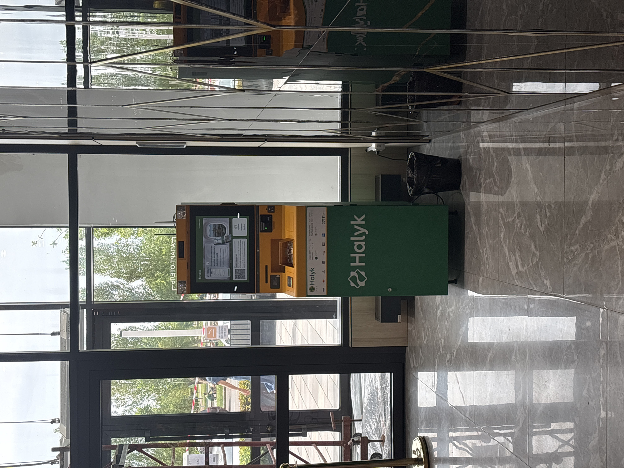 An outdoor ATM machine is shown in front of a building with large glass windows. The ATM is green and yellow, with the Halyk bank logo visible. There are trash cans next to the ATM, and the floor is made of marble tiles. The background shows trees and a blue sky, indicating it is daytime. The scene is likely in Astana, Kazakhstan, as suggested by the presence of Halyk bank, which is a major bank in the country.