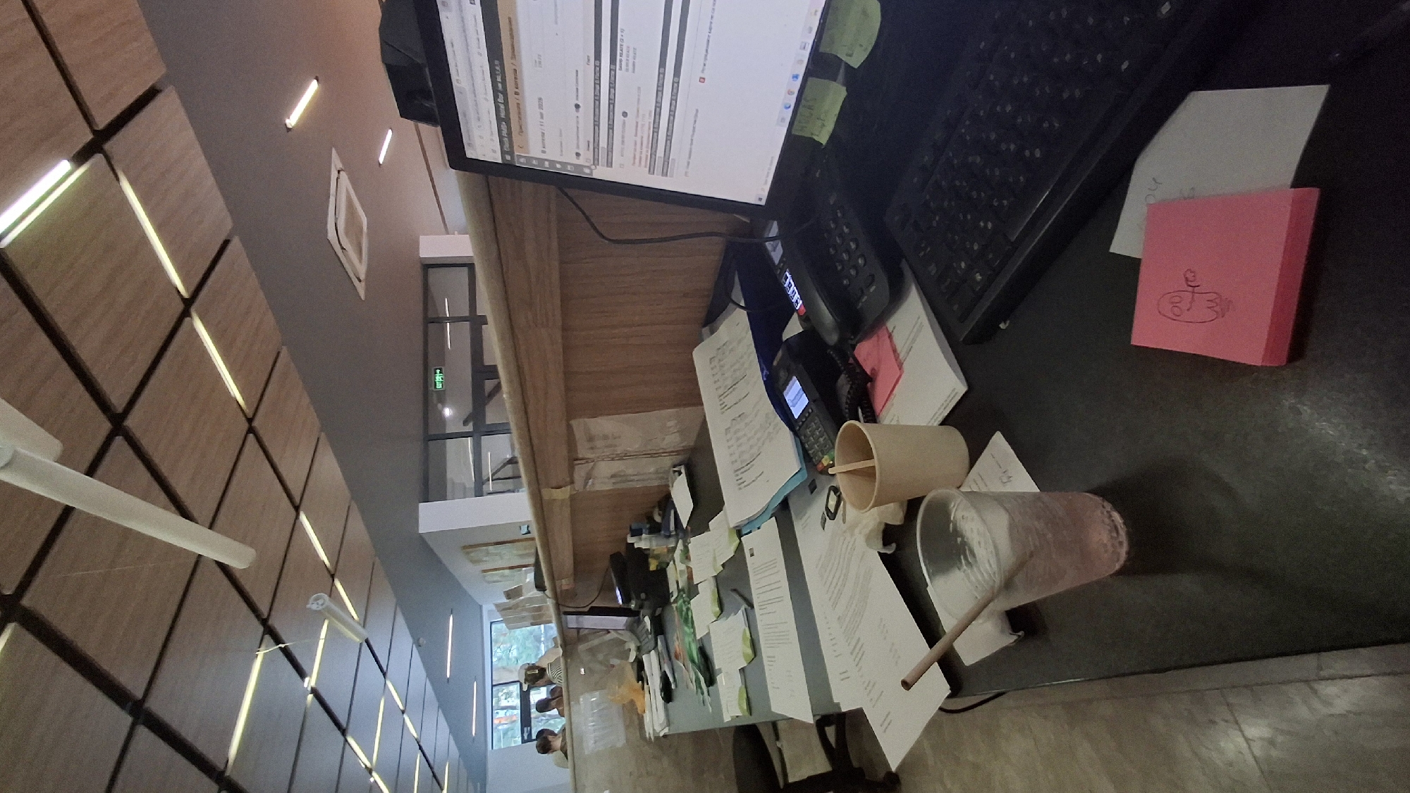 The image depicts a cluttered desk in what appears to be an office or administrative setting in Nesebar, Bulgaria. The desk is covered with various documents, papers, and office supplies. Prominently featured is a laptop with an open web page, suggesting work in progress. To the right of the laptop, a black keyboard is visible. In the foreground, a clear plastic cup filled with a pink beverage and a straw sits on the desk. Adjacent to the cup, a light brown paper cup is positioned near a stack of papers. A pink sticky note with a hand-drawn cartoon of a ghost-like figure is placed on the desk. Several payment terminals are also visible on the desk, indicating a potential service or transaction area. In the background, there is a wooden counter or partition. Beyond the partition, blurry figures of people can be seen, and a sign with "EXIT" in a foreign language and an arrow is displayed on a wall with glass windows. The ceiling features a modern design with wooden panels and integrated lighting. The overall impression is one of a busy workspace with ongoing administrative tasks.