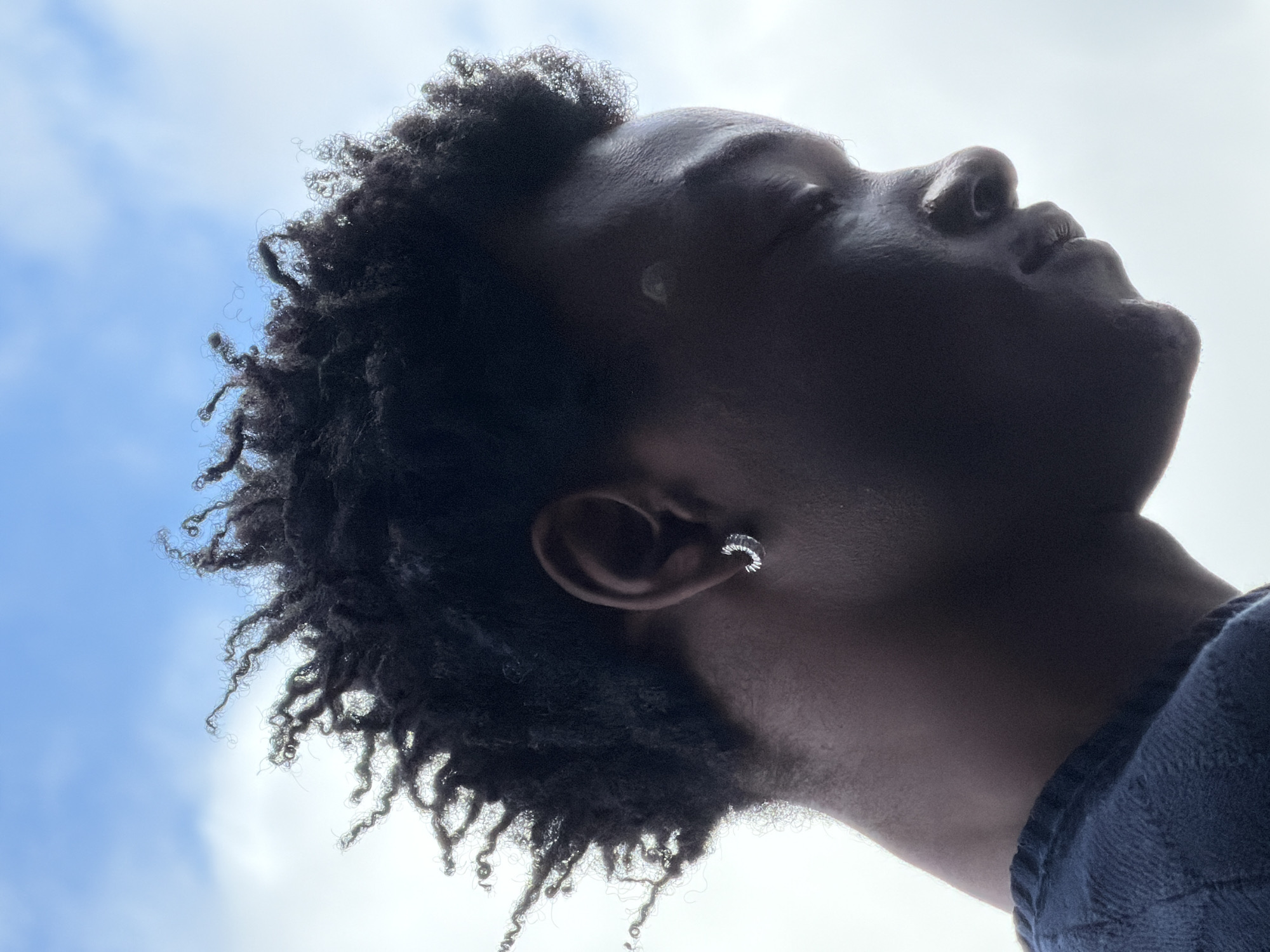 A low-angle, close-up shot depicts a person with dark skin and short, dark, curly hair. The person is looking upwards towards a bright sky with scattered clouds. An earring is visible on their left ear. The person is wearing a dark-colored shirt or top. The lighting creates strong shadows on the person's face and neck. The background consists of a cloudy sky with some visible blue patches. There is no specific information available in the image to determine if this image was taken in London, United Kingdom.