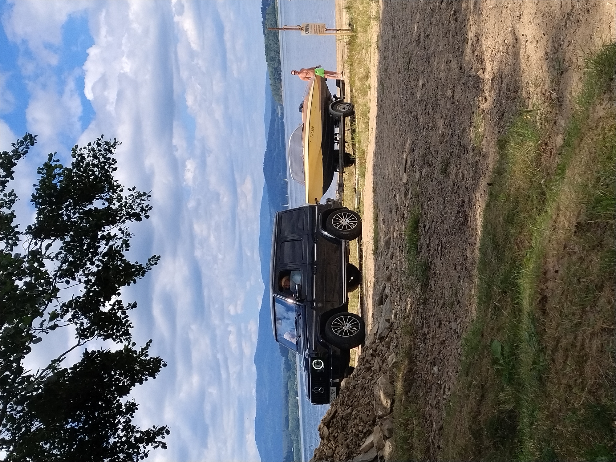 A black Mercedes-Benz G-Class SUV is parked on a dirt path near a body of water. The SUV is hitched to a boat trailer carrying a yellow and brown speedboat. A shirtless man wearing green swim trunks stands beside the boat. In the background, a calm lake is visible with rolling hills and a cloudy sky. A sign that reads "CAMPING" is posted on a wooden pole near the water's edge. The scene appears to be a recreational area, likely at a lake.