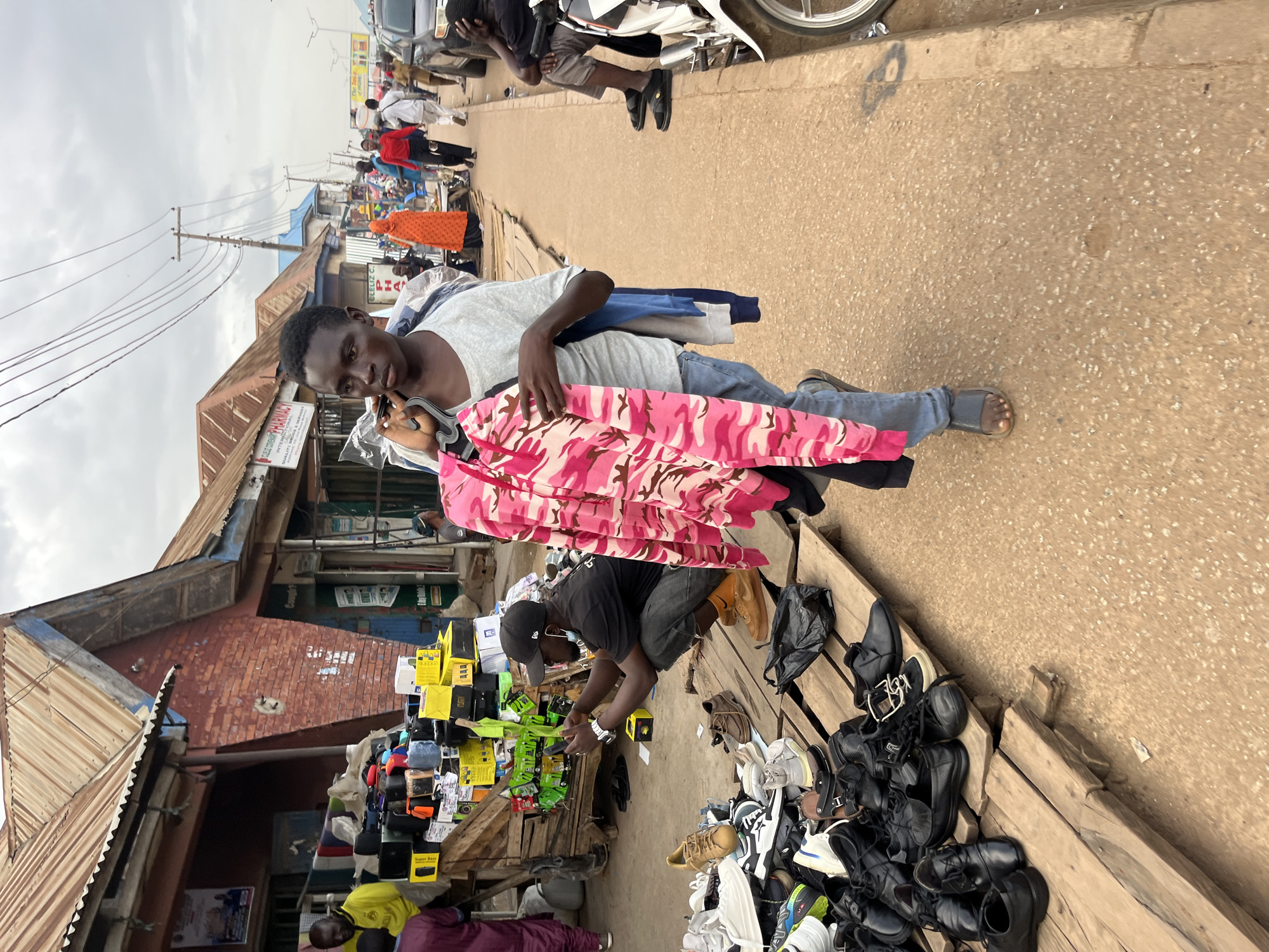 This image depicts a street scene in Bukuru, Nigeria, likely a market area. A young Black male, wearing a light grey t-shirt and blue jeans, walks towards the right of the frame, carrying a pink camouflage-patterned jacket on a hanger over his left shoulder, along with other clothing items. He is wearing dark grey slide sandals. In the background to his left, a Black male wearing a black t-shirt, black baseball cap, and dark pants is crouched down, looking at his phone. He wears a watch on his left wrist and orange socks. In front of him, various items, including stacks of boxes, electronics, and bags, are displayed on a wooden platform. To the right of the walking male, several people are visible in the background, some walking along the street. A motorcycle is partially visible on the far right. The environment appears to be a commercial street with several small shops and stalls lining the left side of the image, featuring corrugated metal roofs. The ground is paved with asphalt, and power lines are strung overhead. The sky is overcast with clouds.