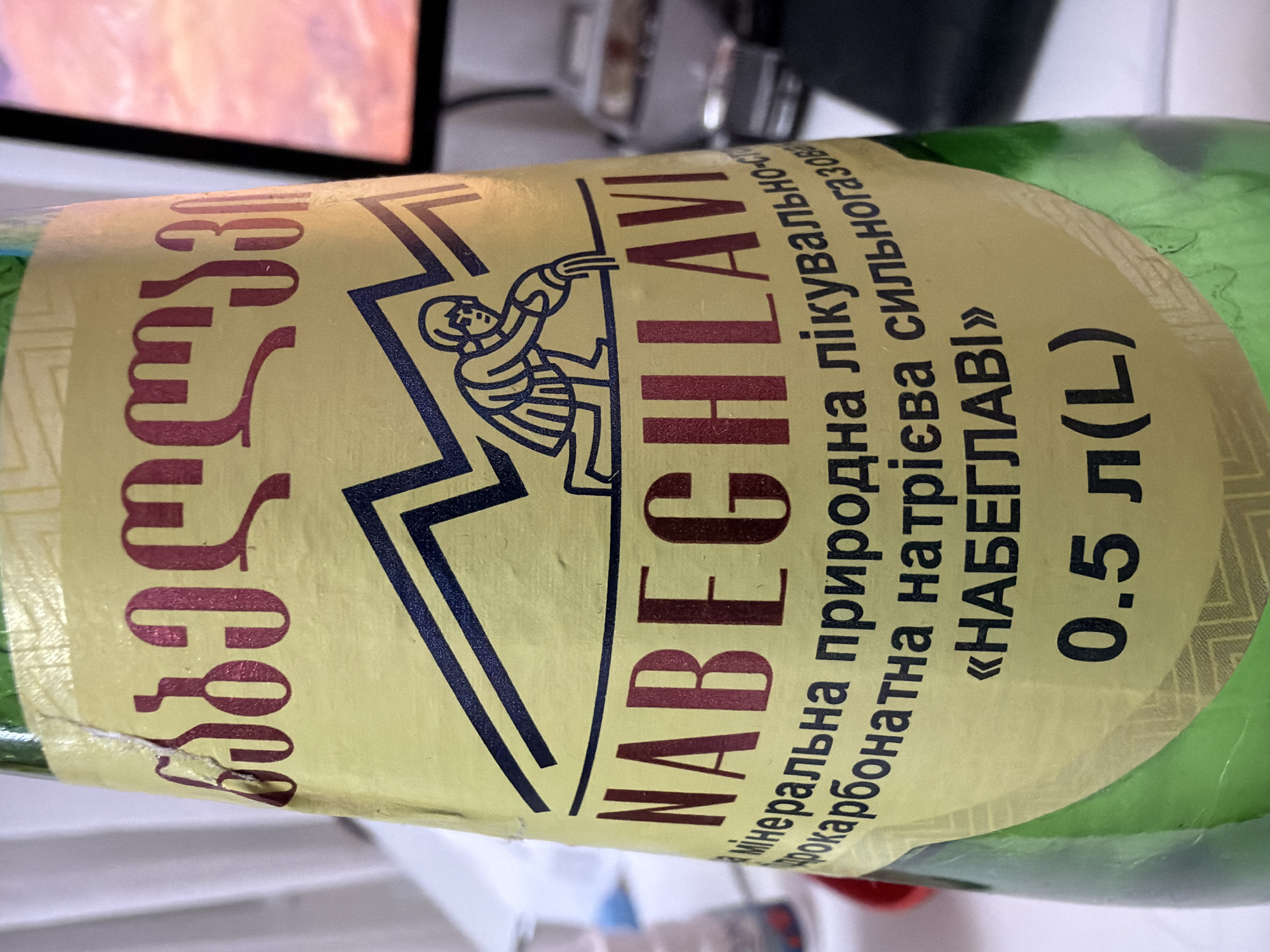 The image shows a close-up of a green glass bottle of water, likely Nabeghlavi, due to the prominent branding. The label is yellow and features text in both Georgian and Cyrillic script, as well as a graphic of a person pouring water. The bottle is sitting on a white surface, and in the background, a computer monitor displays a colorful, possibly sunset-like image. The scene appears to be indoors, in an office or home environment. There is no indication of the location being specifically in Kyiv, Ukraine, beyond the context provided in the prompt.