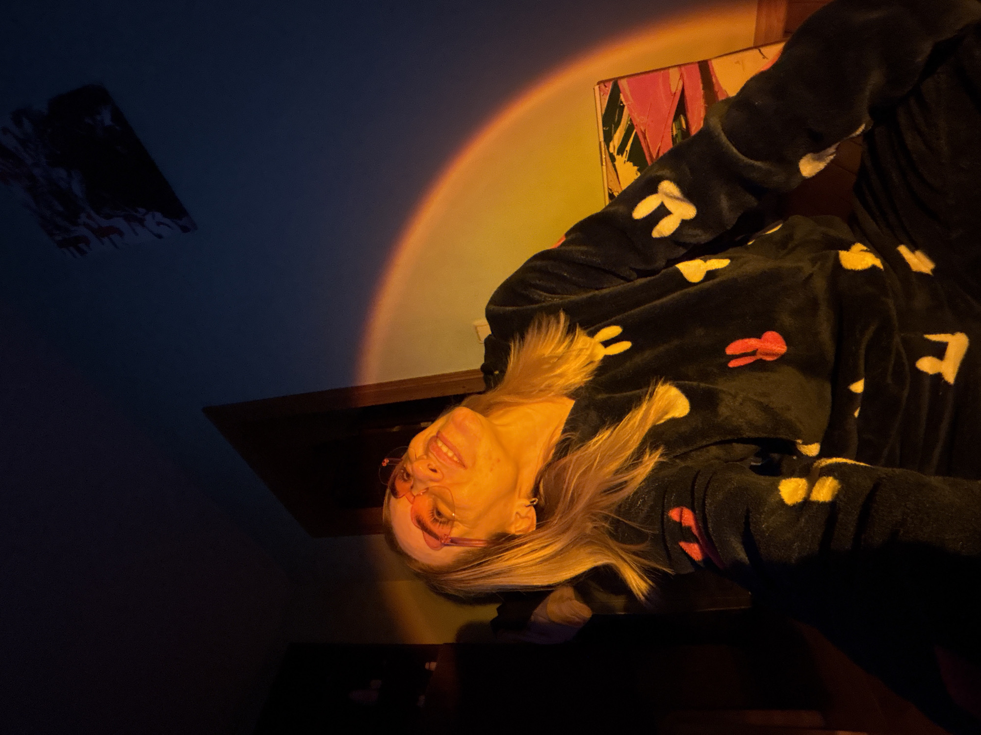 The image shows a woman with blonde hair and pink glasses smiling, wearing a black jumpsuit with red, yellow, and white bunny ear-shaped patterns. The room features a gradient of color: a dark blue transitioning into a golden yellow backdrop resembling a sunset projection. On the left is a piece of artwork hanging on the wall, and another piece is visible above the woman. A dark shelf or table is also apparent in the scene. The location is Unknown, Unknown.
