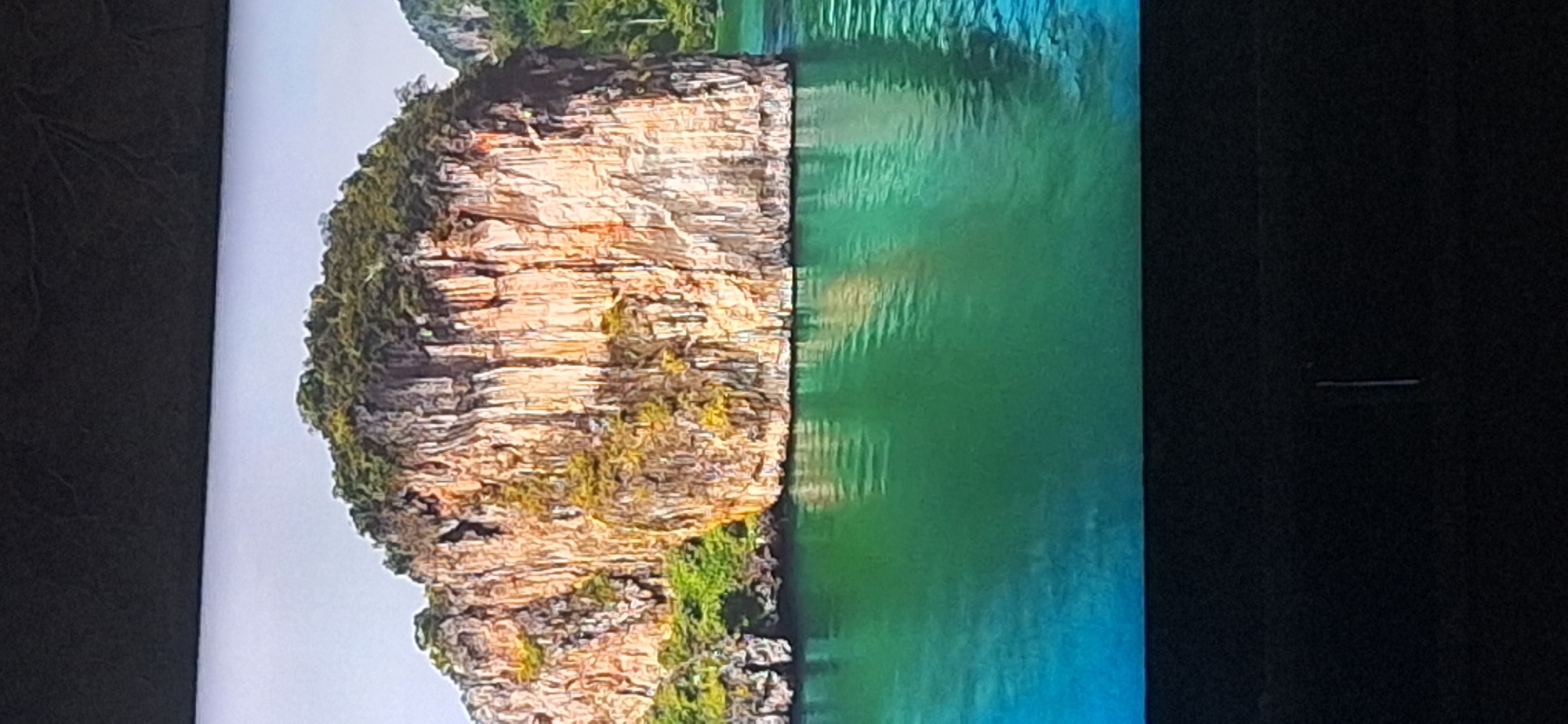 The image shows a rocky island with green vegetation on top, emerging from a body of bright green and blue water. The island's rock face is sheer and appears weathered, with vertical striations. The water is calm, with reflections visible on its surface. The scene is a natural landscape, likely a tropical or sub-tropical location, and has no discernible connection to the city of Bila Tserkva, Ukraine, which is an inland city with no coastal features.