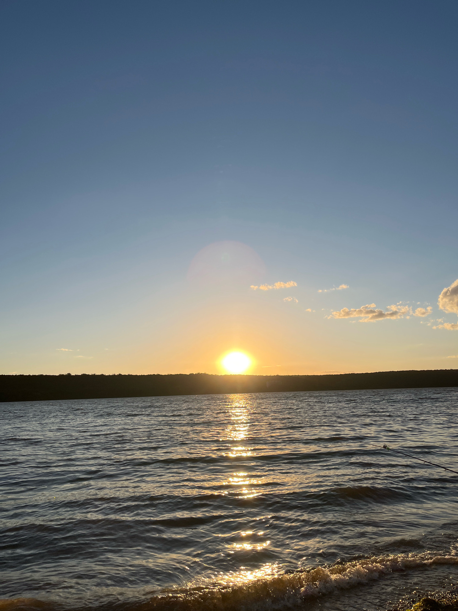 A serene sunset over the Dniester River in Korzhevo, Moldova, casts a warm golden glow on the water. The sun, a bright orb, is partially obscured by a distant, dark treeline that stretches across the horizon. The sky above is a clear, gradient blue, transitioning from a lighter hue near the horizon to a deeper shade overhead, with a few wispy clouds scattered across the expanse. The river's surface is rippled, reflecting the sun's brilliance in a shimmering path of light that leads towards the foreground. Small waves gently lap at a pebble-strewn shore. A fishing rod, propped on the bank, suggests recreational activity. No people are visible in the frame.