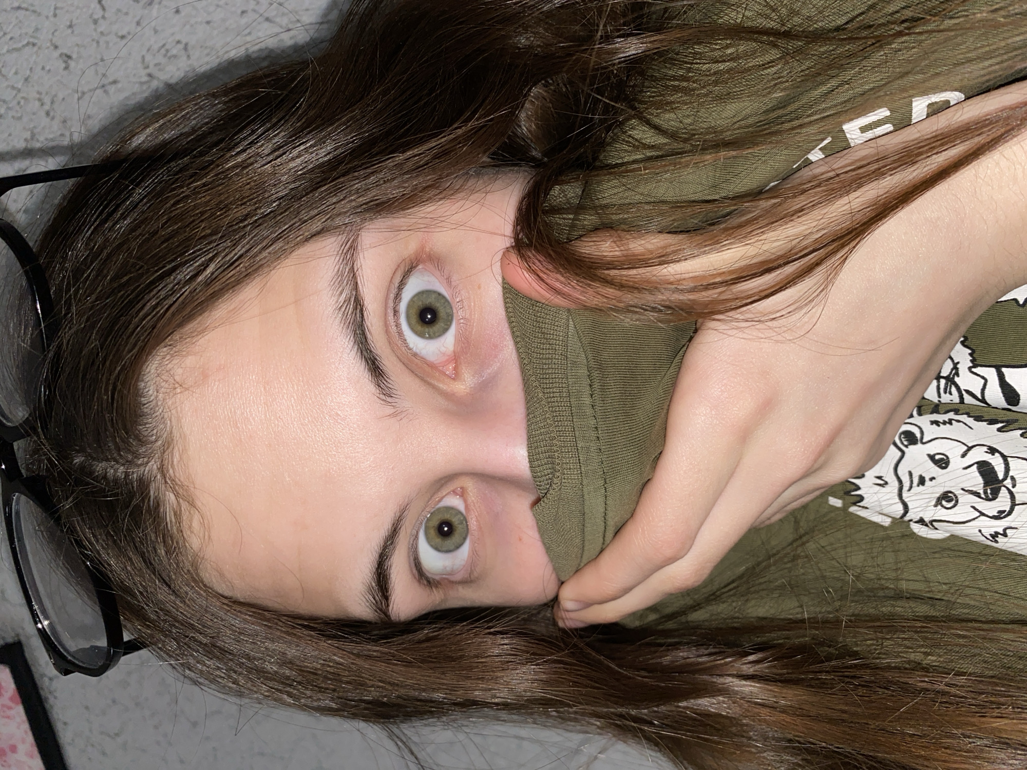 A person with long brown hair, wearing black-rimmed glasses, has pulled up the collar of a green t-shirt to cover their mouth. Their eyes are wide. The background appears to be a gray wall. There is no evidence of the location being Votkinsk, Russia.