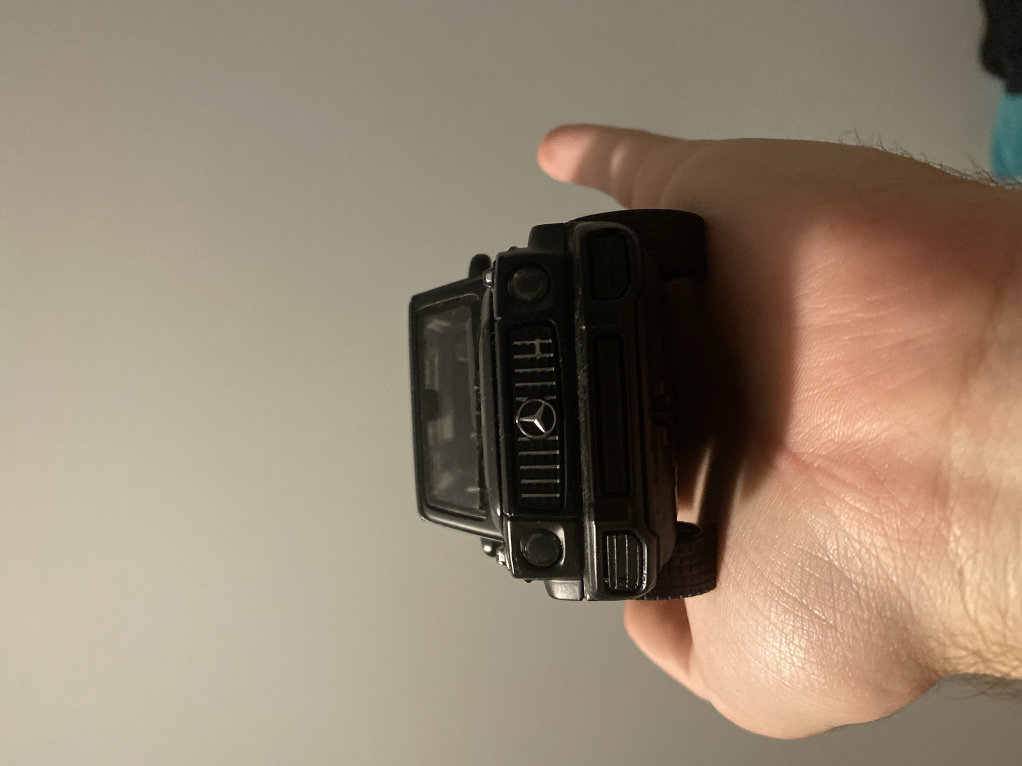 A black toy Mercedes-Benz G-Class SUV is being held in a person's palm, facing the camera. The toy car has a detailed front grill with the Mercedes-Benz logo in the center, and circular headlights. The person's hand is lightly cupped, with the car resting on the palm. The background is a plain, light gray wall. The image does not contain any specific details that would allow for its location to be identified within the city of Łódź, Poland.
