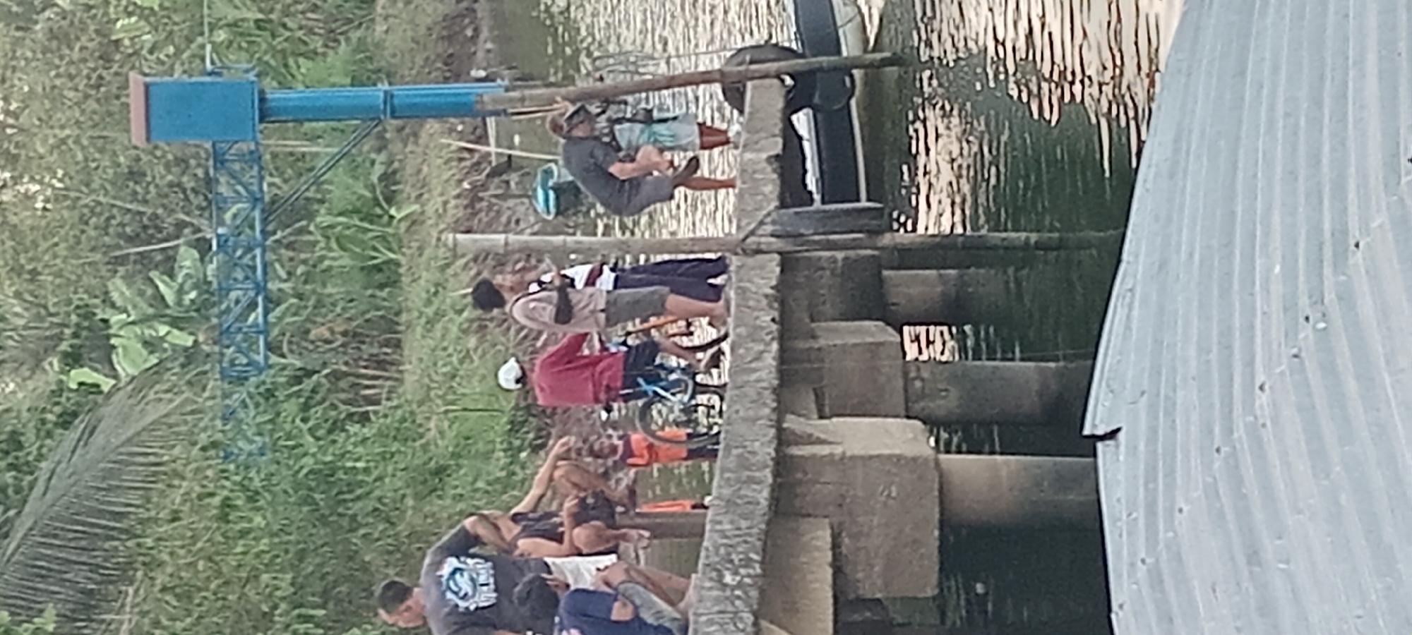 In Kalipucang, Indonesia, a group of individuals are gathered on a concrete pier by a body of water. On the left, a man wearing a dark t-shirt with a graphic print stands on the pier. Next to him, a person is seated, looking down, while another individual in a blue shirt is crouched on the edge. Further to the right, a young child in an orange shirt stands near a bicycle with blue accents. Several other people are standing on the pier, including one in a red shirt, another in a beige shirt, and two in striped and dark shirts. A man wearing a dark shirt and goggles is bending down, possibly adjusting something on his footwear, near a pole. Behind the people, lush green vegetation covers the background. A blue structure with a boxy top is visible in the distance, mounted on a pole with a connecting walkway. A boat is docked alongside the pier. The water surface exhibits reflections of the surroundings. In the foreground, a corrugated metal roof is partially visible.