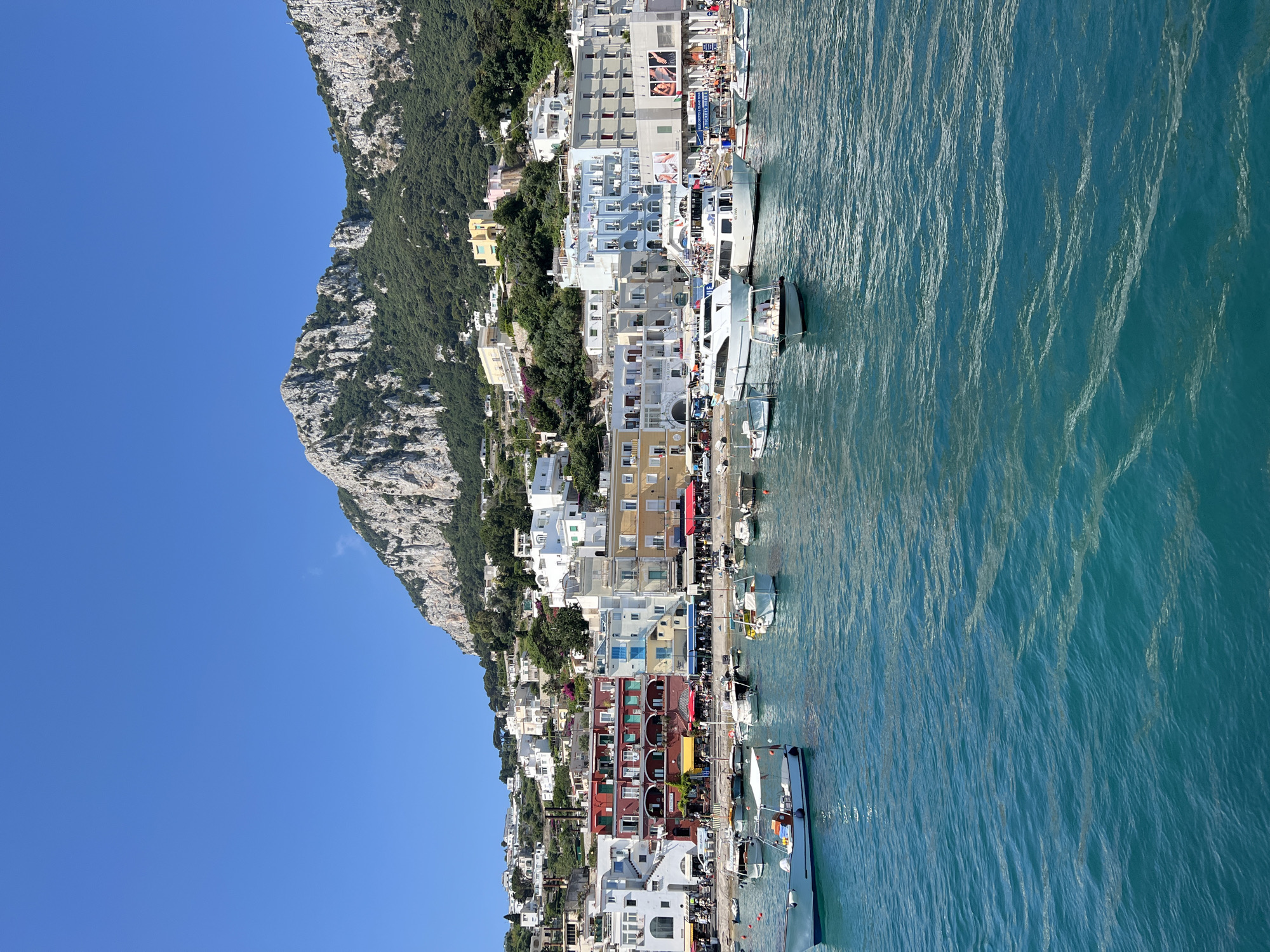 The image shows a view of the Marina Grande in Capri, Italy. The foreground features clear blue water with several boats anchored, ranging from small dinghies to larger yachts. In the midground, a colorful waterfront of buildings is visible, housing shops and restaurants with awnings. These buildings are painted in various shades of white, yellow, blue, and red, creating a vibrant facade. Behind the waterfront, the town of Capri rises up the hillside, characterized by its dense collection of white buildings with terracotta roofs. The background is dominated by a dramatic, rocky mountain range covered in lush green vegetation, with a clear blue sky overhead. People are visible on the waterfront promenade and on some of the boats, suggesting a lively atmosphere.