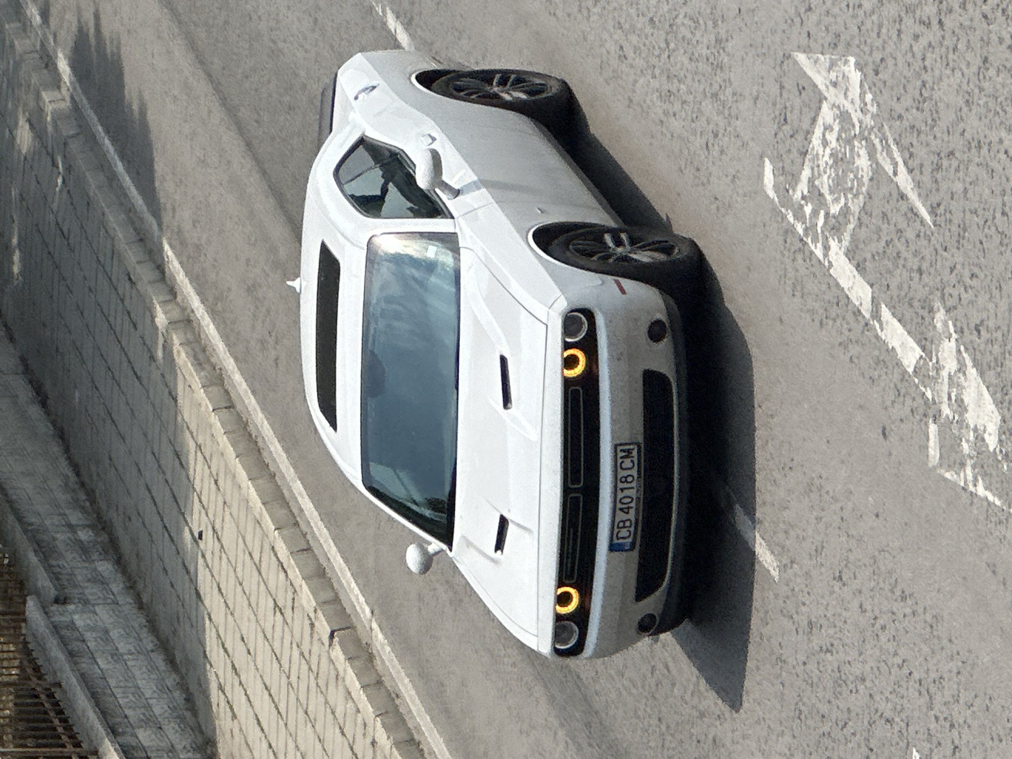 A white Dodge Challenger is parked on a paved road next to a concrete retaining wall. The car is viewed from a high angle, tilted to its side. The front of the car is facing towards the viewer's left, and its license plate reads "CB 4018 CM". The road has painted white markings, including a sharp zig-zag pattern, possibly indicating a turning lane or a hazard. The retaining wall is made of rectangular concrete blocks and has a textured surface. The scene appears to be outdoors during the daytime, with sunlight casting shadows on the road and wall. The sky is not visible in the frame, but the bright lighting suggests a clear day. There are no people visible in the image. The setting suggests an urban or suburban environment, possibly on a street or a parking area.