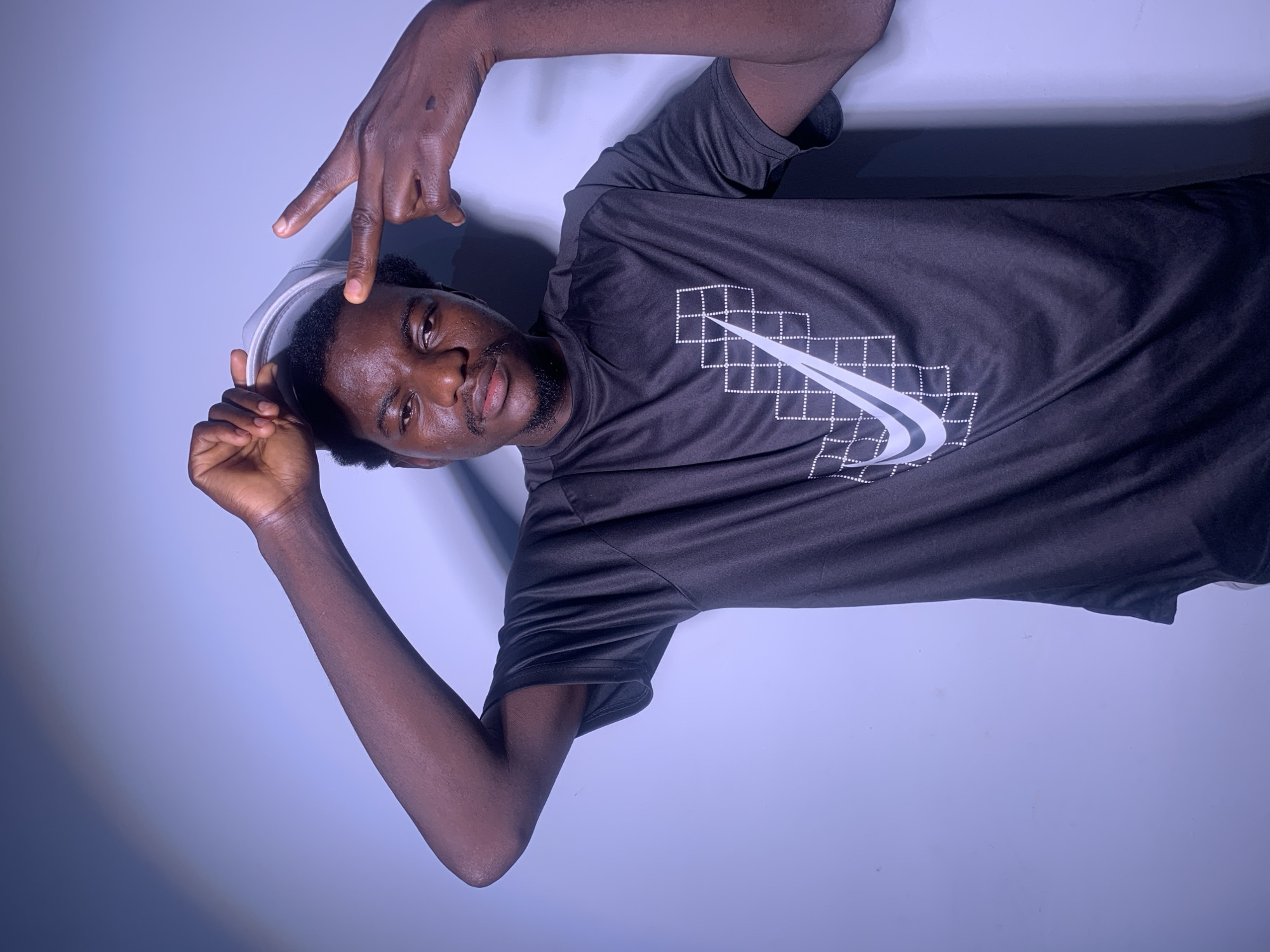 A young Black man with dark skin and curly black hair is posing against a plain, light purple wall in a studio setting. He is wearing a short-sleeved black t-shirt with a white Nike logo on it. His left arm is raised, and he is holding a light gray baseball cap on top of his head with his left hand. His right arm is also raised, and his index and middle fingers are extended, pointing towards his own head. He has a faint goatee and mustache. The lighting creates a shadow on the wall behind him. The image appears to be a headshot or portrait. The location is not specified.