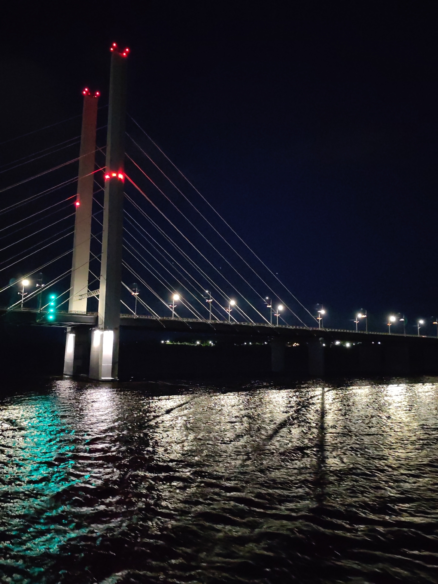 A nighttime shot captures a cable-stayed bridge spanning a body of water. The bridge features two tall, light-colored towers with red lights at their peaks and lower points. Cables extend from these towers to support the bridge's roadway, which is illuminated by evenly spaced streetlights. The water reflects light from the bridge and surrounding structures, creating a shimmering effect. The background is dark, with faint lights visible on the distant shore. The location is in the city of Unknown, Unknown.