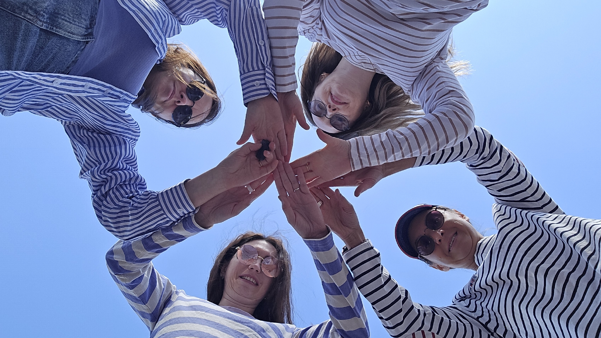 A low-angle, full-frame shot captures four individuals from the chest up, their arms extended upwards and hands clasped in the center against a clear blue sky. The subjects are seen from below, creating a sense of looking up at them. Each person is wearing a horizontally striped long-sleeved shirt. From clockwise, starting at the top: a person in a blue and white striped shirt wears sunglasses and has long brown hair. To their right, a person in a blue and white striped shirt also wears sunglasses and has brown hair. Below them, a person in a white and brown striped shirt is wearing sunglasses. Finally, at the bottom left, a person wearing a red and black baseball cap and sunglasses is in a black and white striped shirt. The central gathering of hands suggests a moment of unity or a shared activity, with rings visible on several fingers. The overall impression is one of camaraderie and outdoor activity under a bright sky. There is no identifiable information in the image that would place this scene specifically in Chaikovsky, Russia.