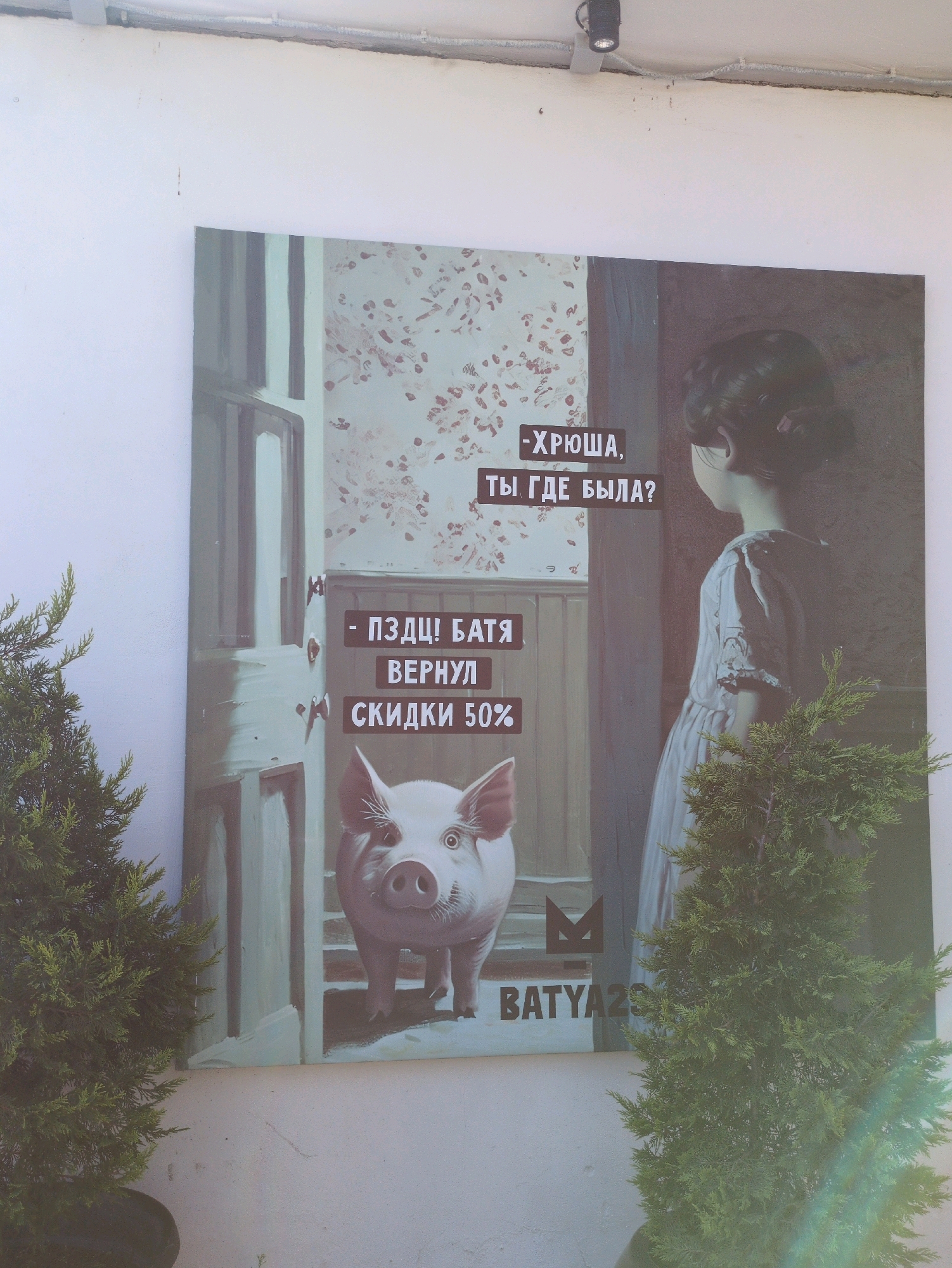 An outdoor, eye-level, medium shot captures a large painting mounted on a white wall, partially obscured by two small green evergreen trees in planters. The painting depicts a scene with a young girl in a light-colored dress, her back turned to the viewer, her eyes covered by her hair. Beside her, a pig's face is visible, looking directly at the viewer. Text overlays are present on the painting. In the upper right corner, text reads "-ХРЮША, ТЫ ГДЕ БЫЛА?". In the lower left section, text reads "-ПЗДЦ! БАТЯ ВЕРНУЛ СКИДКИ 50%". A stylized "M" logo is visible above the text "BATYA29" in the lower right portion of the painting. The wall is textured and appears weathered. Electrical conduits and a spotlight are mounted on the ceiling above the painting.