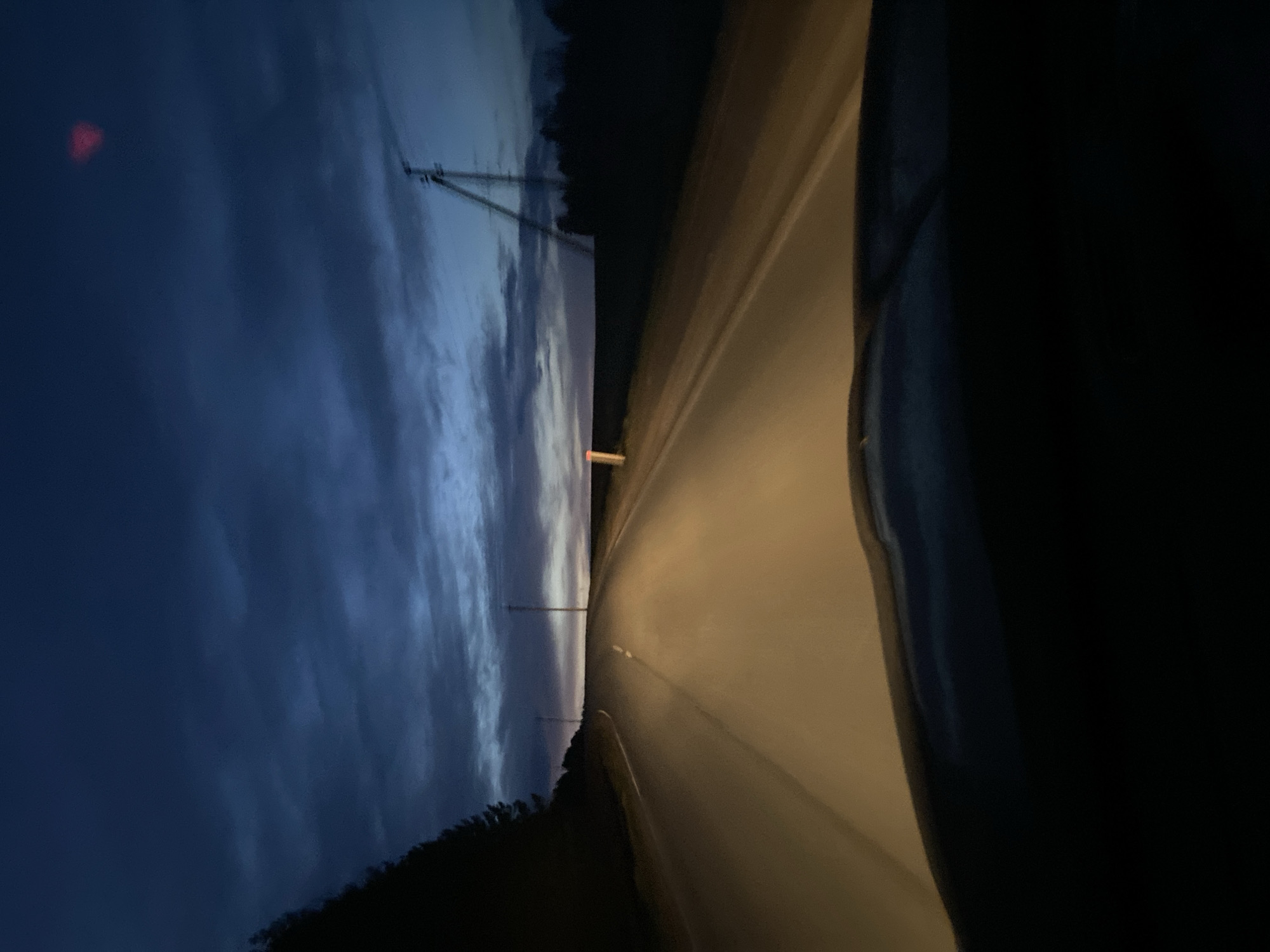 The image captures a twilight or nighttime scene from the interior of a vehicle, looking out onto a road in Kamennyi Kliuch, Russia. The sky, a deep indigo blue with soft, streaky cloud formations, dominates the upper-left and left side of the frame. A small, distinct red light, possibly a distant tower light or an external reflection, glows in the upper-left corner of the sky.
The road occupies the right side of the image, brightly illuminated by a warm, yellowish light, suggesting the vehicle's headlights. It appears to curve gently as it recedes into the distance, meeting a dark, indistinct horizon line formed by trees or low hills. Several slender utility poles or communication towers are silhouetted against the lighter part of the sky, with one pole featuring a prominent triangular support structure.
The extreme right side and lower portion of the image are very dark and out of focus, indicating the presence of the vehicle's interior, such as a dashboard or door frame. No people are visible, nor is any discernible text. The overall scene evokes a sense of tranquil travel through a rural or semi-rural landscape during the early morning or late evening hours.