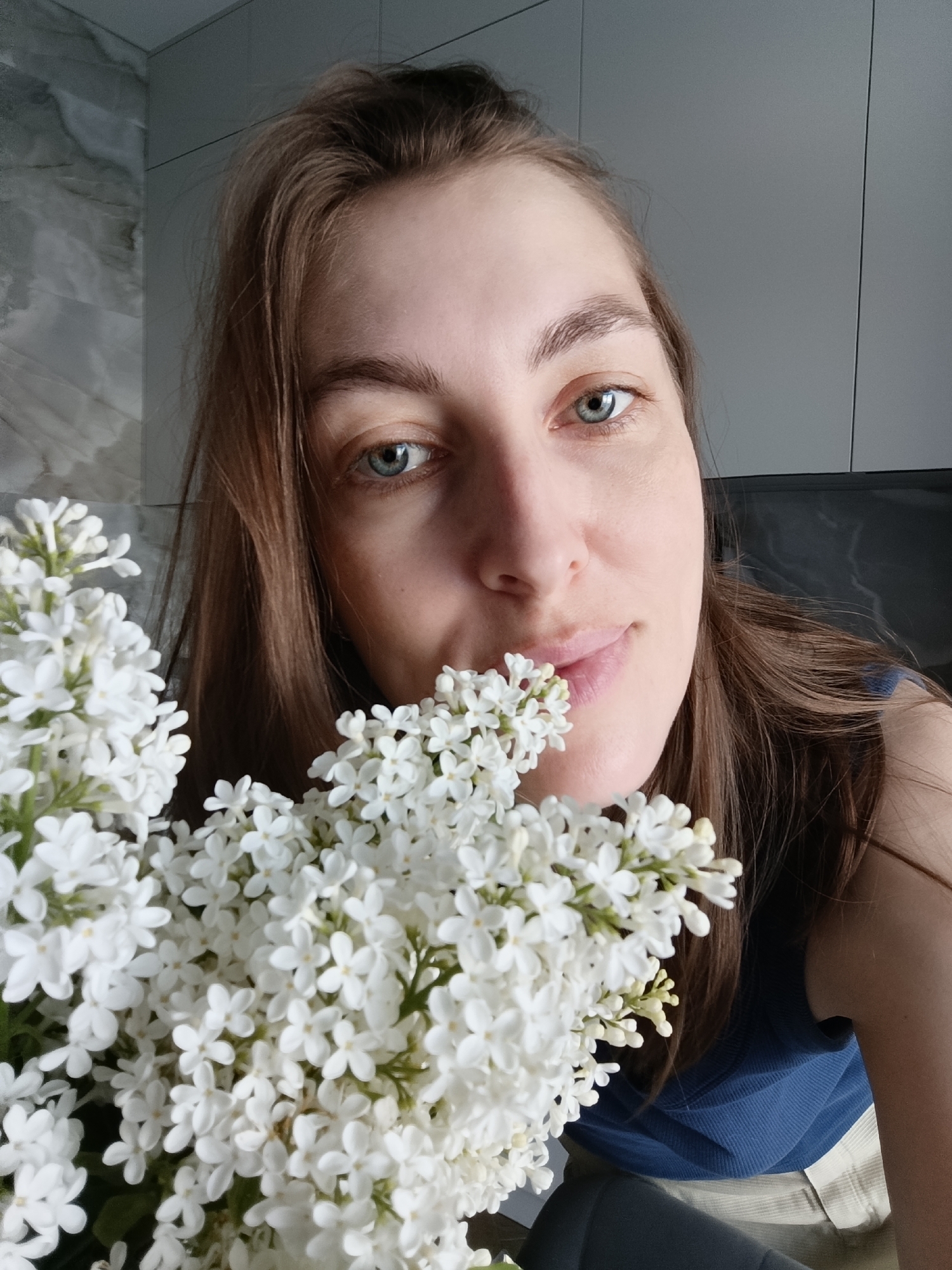 A woman with light brown hair and blue eyes is posing with a bouquet of white flowers. The woman is wearing a blue shirt. The location appears to be indoors, possibly a home, with a marble-patterned wall and gray cabinets visible in the background. There is no information to confirm that the location is Chaikovsky, Russia.