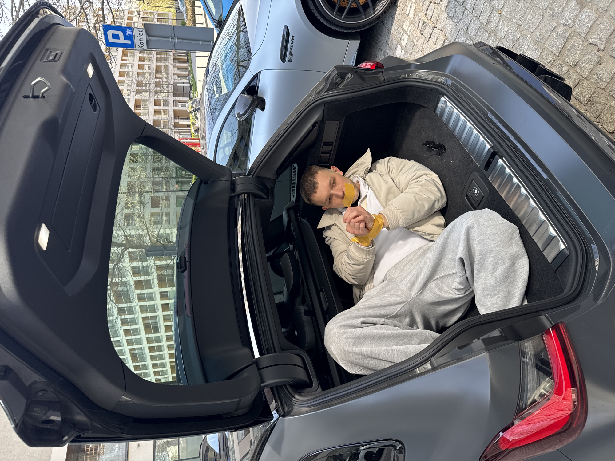 A man with yellow tape over his mouth and around his wrists is lying in the trunk of a gray hatchback vehicle. The car is parked in Warsaw, Poland, as indicated by the "P Koniec" parking sign visible in the background. A light gray car is parked nearby. The trunk is open, exposing the interior and the man inside. The environment includes buildings and trees, indicating an urban setting.