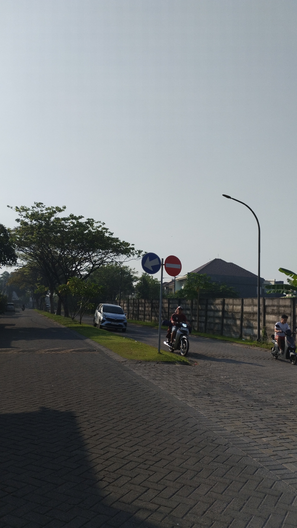 The image depicts a street scene in Sidodadi, Indonesia, under a clear, bright sky. The foreground is dominated by a paved road, likely made of interlocking bricks, with a distinct shadow cast across it from the left. A grassy median runs along the right side of the road.
On the right side of the paved area, two individuals are riding motorcycles. The first motorcycle, closer to the viewer, carries two people; the rider is wearing a dark-colored shirt and a head covering, and the passenger is less clearly visible. The second motorcycle, further down the road, has a single rider wearing a light-colored shirt.
A tall, curved streetlamp stands on the right side of the road, extending into the sky. Two traffic signs are mounted on a single pole. The upper sign is a blue circle with a white arrow pointing left, indicating a mandatory direction. The lower sign is a red circle with a white horizontal bar, signifying a "no entry" or similar prohibition.
To the left of the motorcycles and signs, a silver car is parked on the side of the road, facing away from the viewer. Beyond the car, a row of trees with dense foliage lines the left side of the road, extending into the distance. A concrete wall, likely a boundary for properties, runs along the right side of the road, separating it from buildings that are partially visible behind it. The architectural style of these buildings suggests a residential area. The overall impression is a quiet, possibly suburban street in the morning or late afternoon, given the quality of light and the presence of shadows.