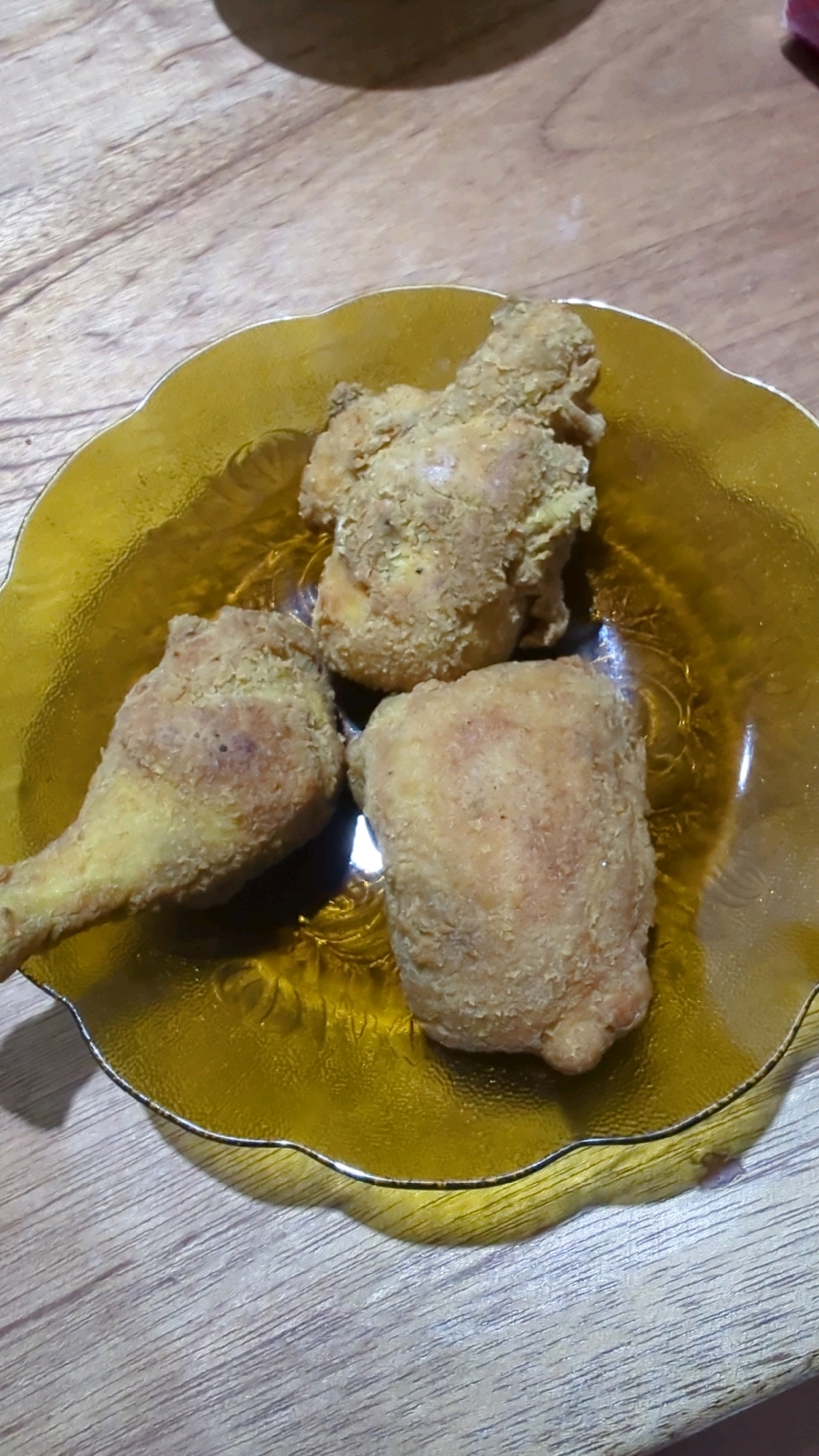 Three pieces of fried chicken are arranged on a translucent amber-colored glass plate. The plate has a scalloped edge and a textured interior pattern. The fried chicken appears golden brown with a crispy coating. The plate rests on a wooden surface, likely a table, with visible wood grain. The lighting suggests an indoor setting, possibly a home. There are no people or any other objects visible in the frame. The location in Baroqah, Indonesia is not discernible from the image itself.
