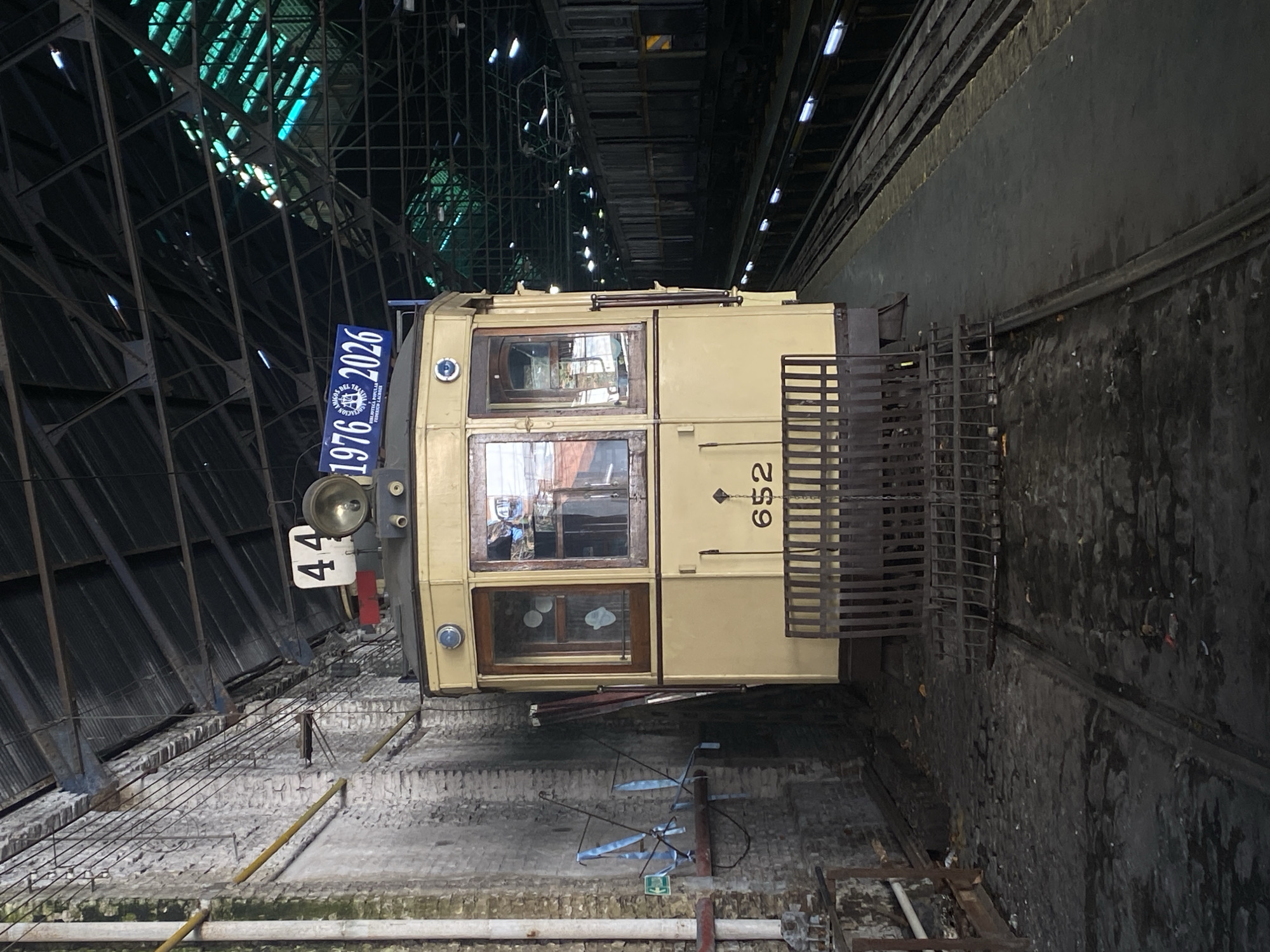 The image shows a vintage railcar, likely a subway car, at the Polvorín Workshop, a restoration and maintenance facility for historic subway cars in Buenos Aires, Argentina. The car is light yellow with brown framed windows and the number 652 on its side. It's situated on tracks within the workshop, which has a high, metal-framed ceiling with greenish illumination. A sign with "1976-2026" is visible on the car's front. The surrounding environment includes brick and concrete walls, and various pipes and conduits associated with the workshop's infrastructure.
