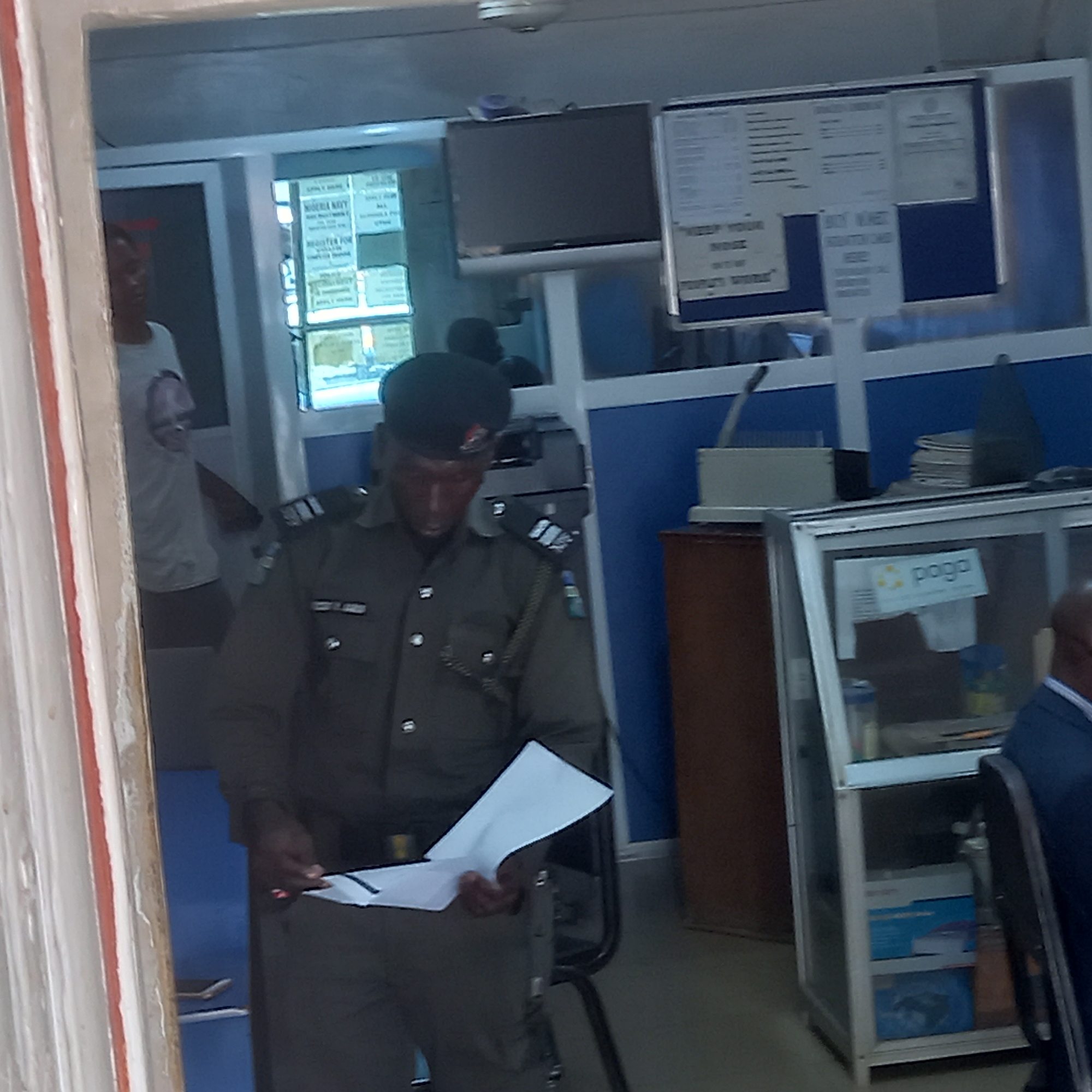 The image depicts an indoor administrative or office setting, likely in Nigeria given the uniform and visible text. In the foreground and center, a man wearing a dark green/khaki uniform, consistent with a Nigerian police or military officer, is standing and engrossed in reading or reviewing several white papers he holds in his hands. He wears a dark cap. Behind him to the left, another person in a white t-shirt is partially visible near a doorway or window. On the far right, a person's head and shoulder, wearing a dark blue garment, are visible.
The scene features typical office elements. A flat-screen television is mounted on the back wall. Below and to its right, a bulletin board displays various documents, some affixed with pins. Visible text on the bulletin board includes "NIGERIA NAVY," "REGISTER FOR," and phrases like "KEEP YOUR" and "SCRATCH CARD HERE." Behind the officer, what appears to be a printer or copier is visible. To the right of the officer, a wooden cabinet or desk section is present, along with a glass display cabinet containing boxes and other items. A prominent "paga" logo and text are visible on a sign within this glass cabinet. The partition walls are light-colored with blue accents. The overall lighting suggests it is daytime. No strong emotions are discernible from the subjects.