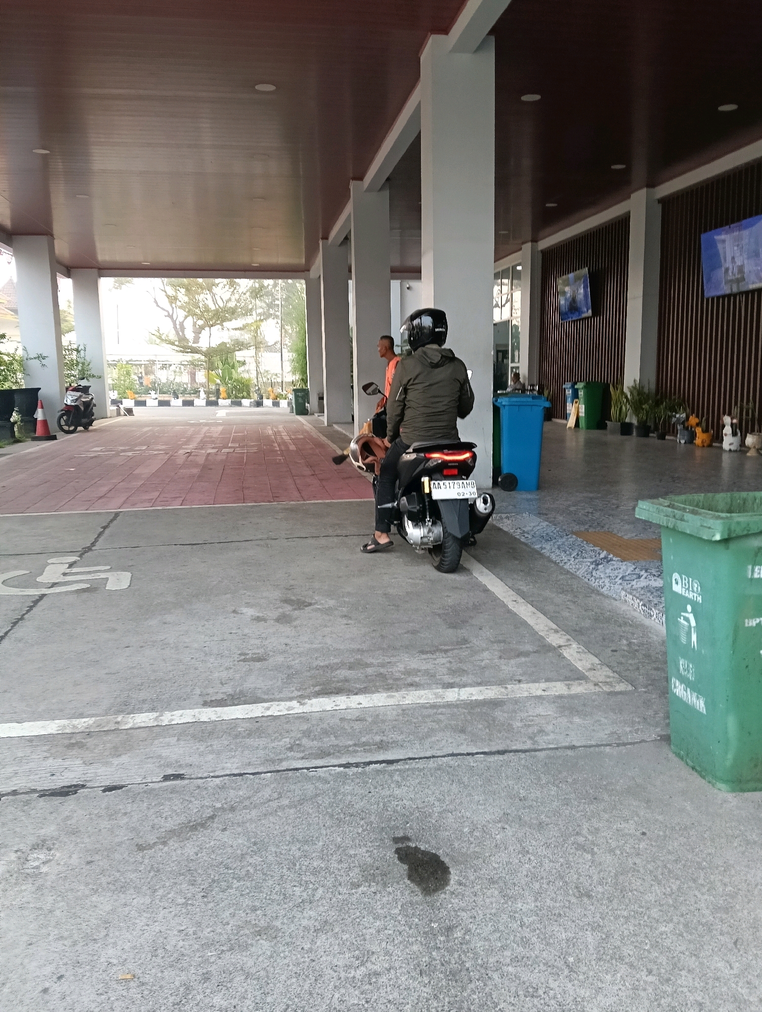 The image depicts a covered walkway or overhang of a building. In the foreground, a concrete surface is marked with white lines, including a disabled parking symbol. A green trash bin labeled "BIO EARTH" and "ORGANIK" is situated on the right side of the frame. In the middle ground, two individuals are seated on a motorcycle. The person in the rear, wearing a black helmet and a dark jacket, is looking away from the camera. The motorcycle has a license plate that reads "AA 5179 AMB" and a date "02.30". To the left of the motorcycle, another motorcycle is parked. Further in the background, the area opens up to a red-paved pathway with several parked cars and trees. In the distance, to the right, a tall wooden-slatted wall is visible with several flat-screen televisions mounted on it. The overall environment suggests an outdoor public space, possibly an entrance or parking area of a commercial or public building. The scene is well-lit, indicating daytime. The context of Magelang, Indonesia, implies that the architecture and potentially the license plate style are characteristic of that region.