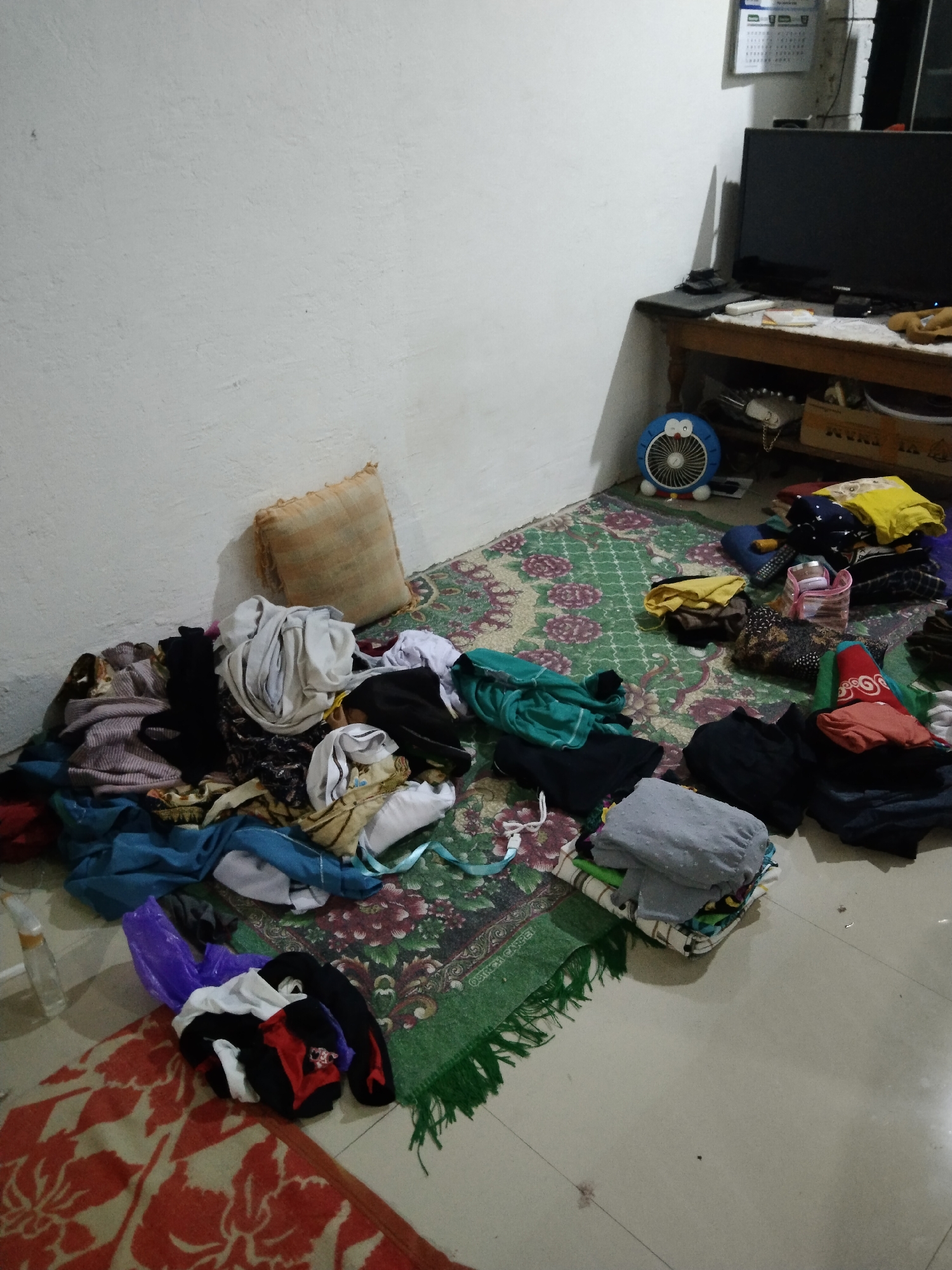 The image depicts a cluttered room, likely a bedroom or living area, with laundry strewn across the floor and on a rug. In the foreground, a pile of folded clothing is stacked on top of other garments. To the left, a plastic bag containing clothes sits on a patterned rug. A larger pile of unorganized laundry is visible further back, partially obscuring a throw pillow. A small blue electric fan with a Doraemon design is positioned on the rug near a wooden table. On the table, a television is visible, along with a calendar hanging on the wall above it. The walls are painted white, and the floor appears to be tiled. The scene suggests a domestic setting in a residential area, potentially in the city of Buntalan, Indonesia, given the context provided. There are no people visible in the image.